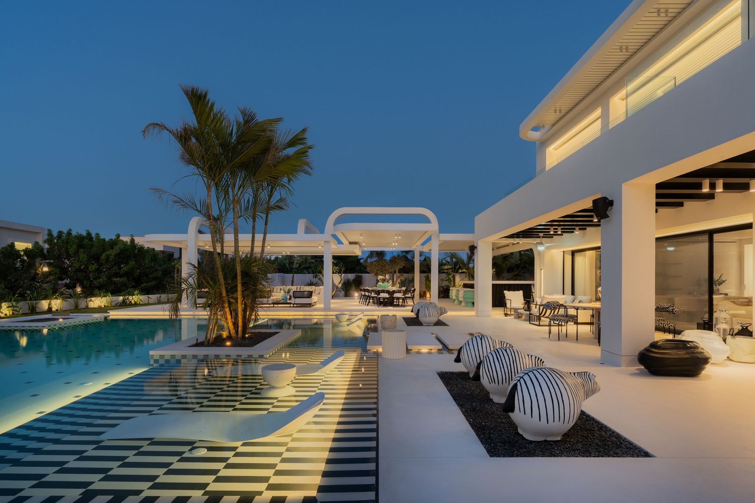 Stunning luxury villa pool deck at dusk with checkered tiles, white pergola, palm trees, and elegant outdoor seating.