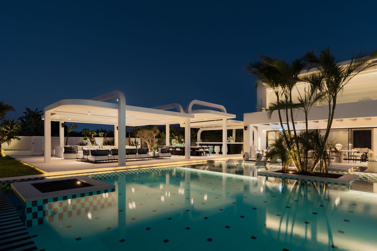 Resort-style white villa with illuminated pool, arched pergola, lush palm trees, and outdoor lounge area at night.