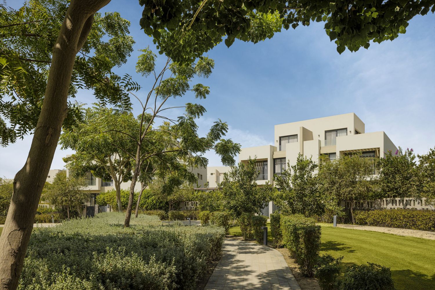 Lush green garden pathway lined with trees and manicured hedges leading to modern white residential villas under blue sky.