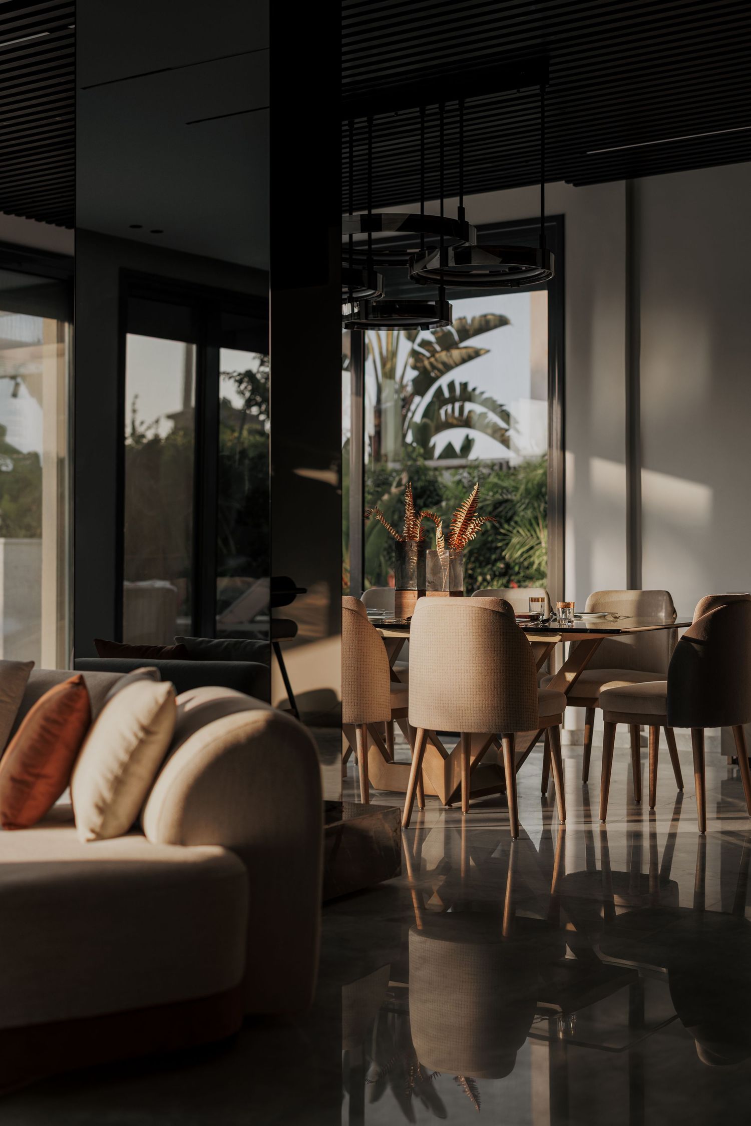 Sunlit dining area with beige chairs, round table, and tropical garden views through floor-to-ceiling windows.