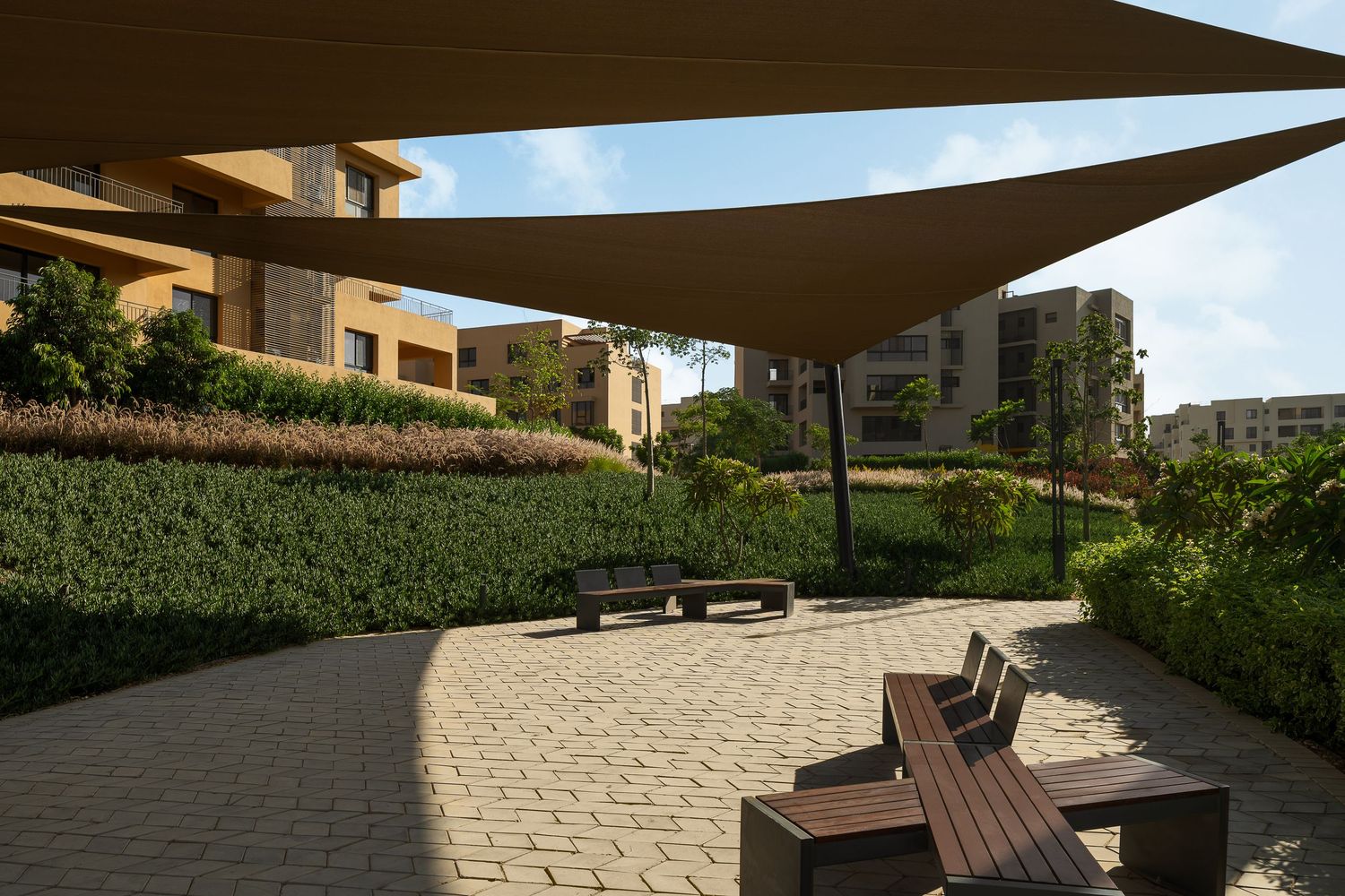 Shaded courtyard seating area with wooden benches under large sail canopies surrounded by lush green hedges.