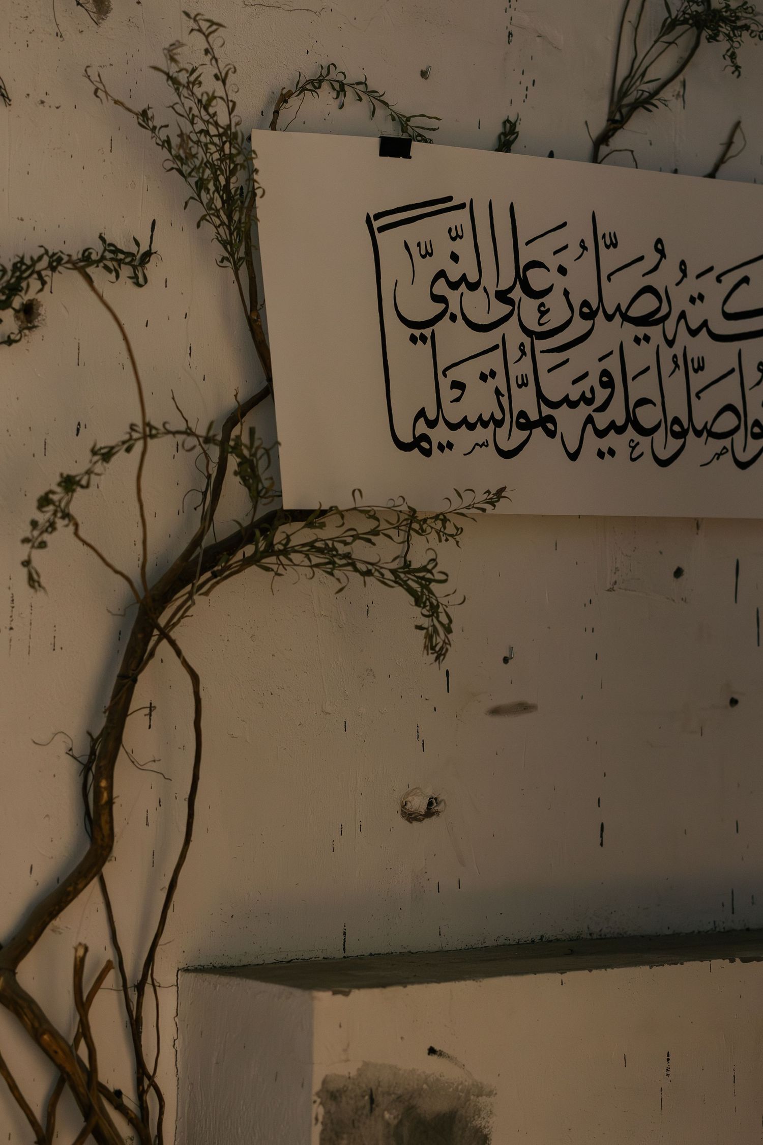 Arabic calligraphy text on white paper mounted on a weathered wall with dried plant branches.