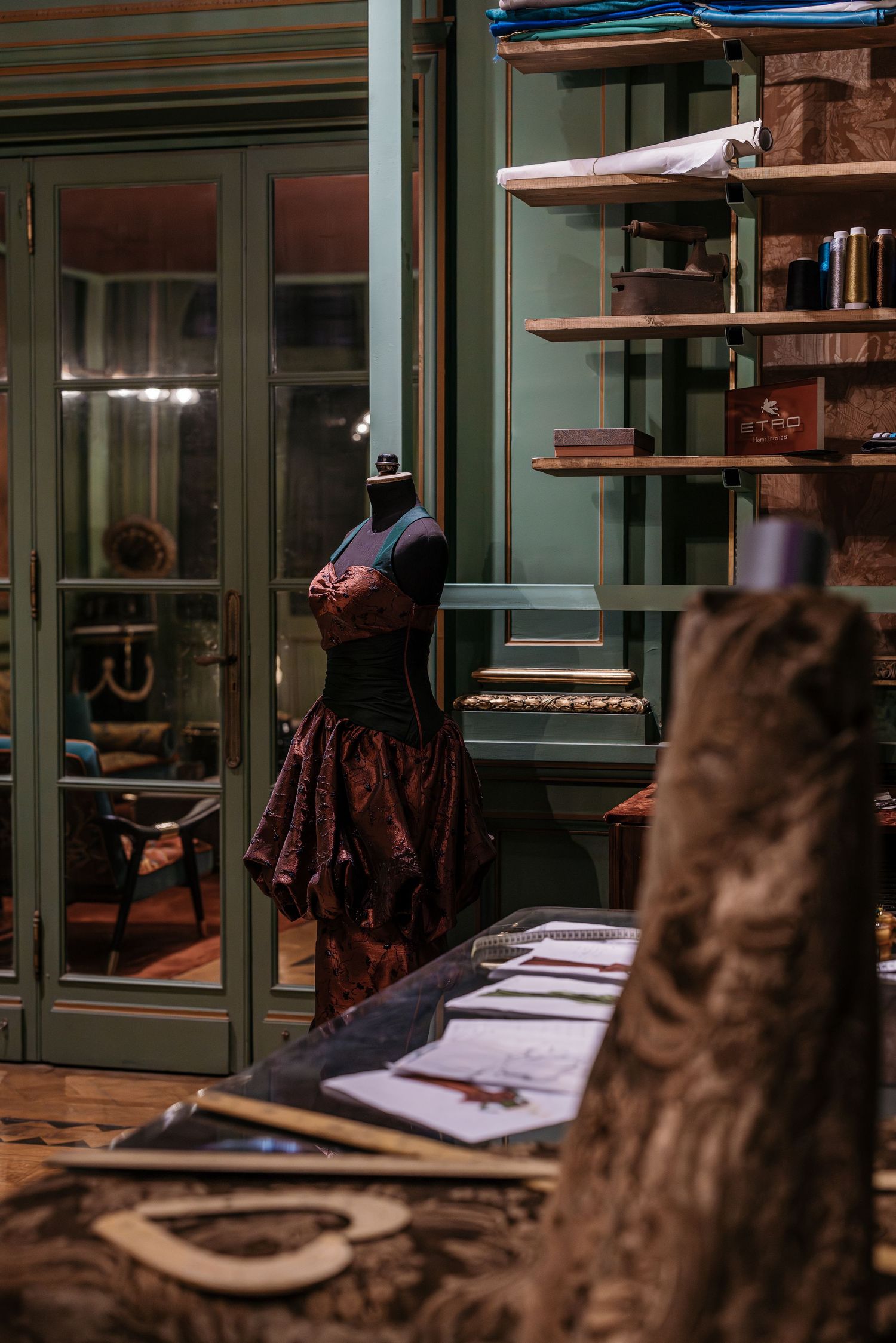 Dressmaker mannequin in ornate corset gown displayed in elegant atelier with green shelving and glass doors.