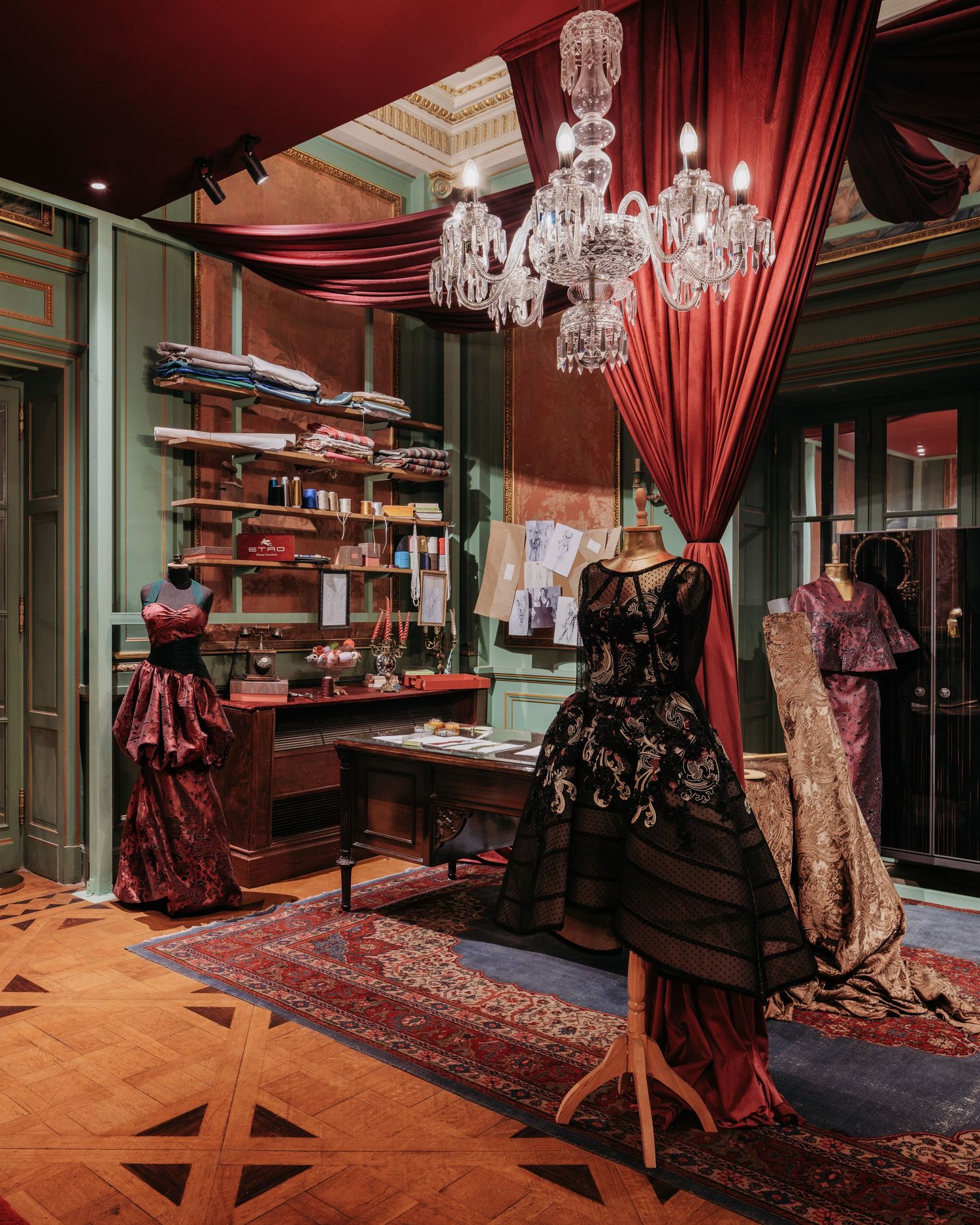 Elegant fashion atelier with ornate gowns on mannequins, crystal chandelier, red drapes, and antique wooden desk.