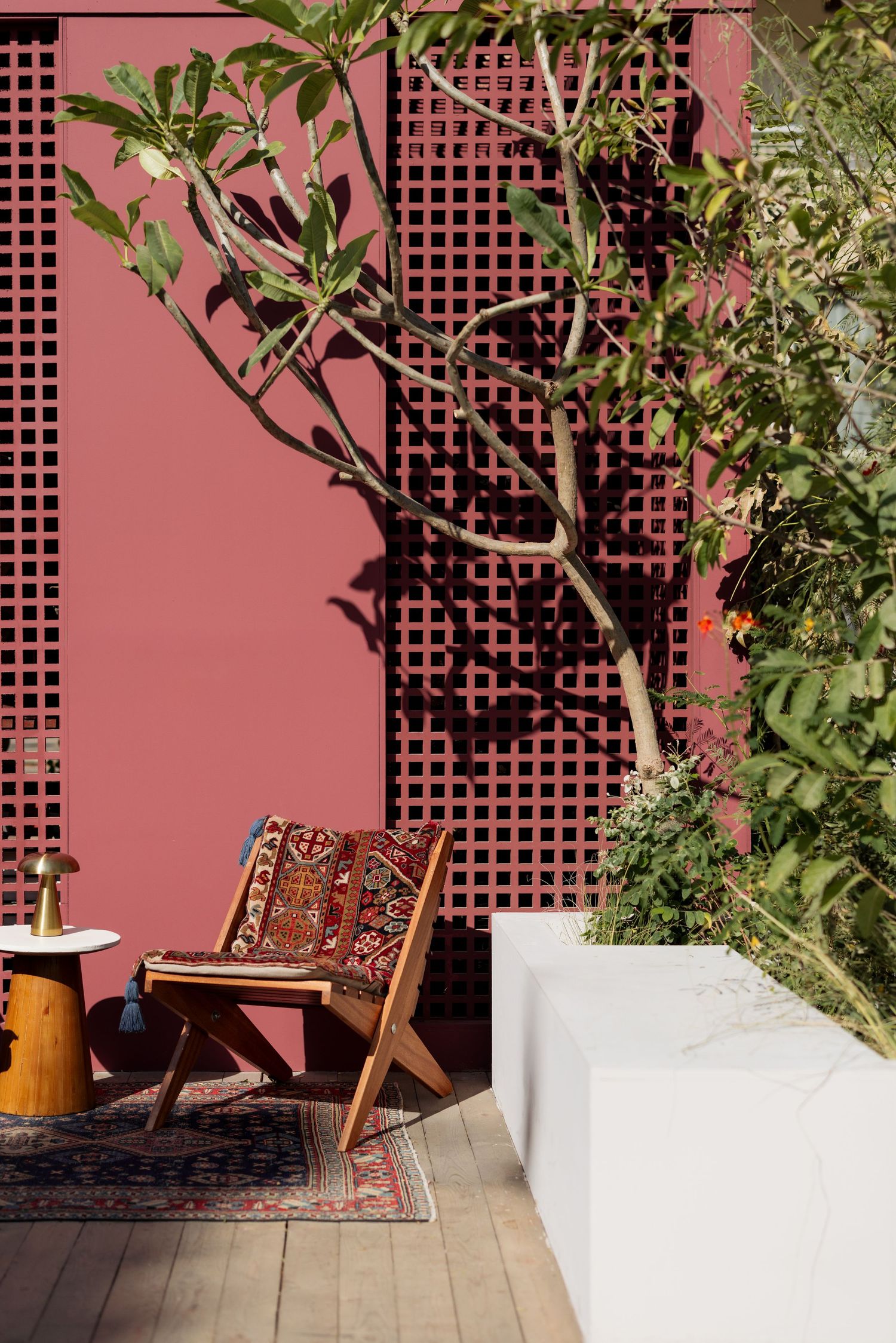 Wooden chair with patterned cushion beside a pink lattice wall with tree shadows in a sunny courtyard.