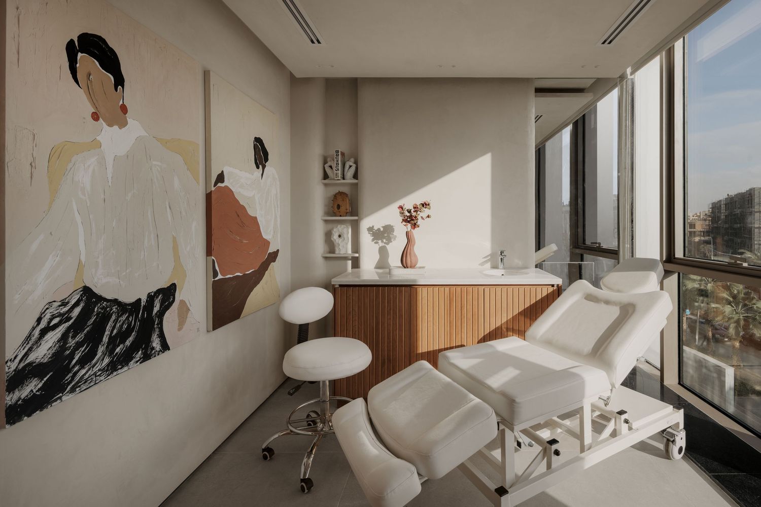 Bright spa treatment room with white massage bed, large artwork, and panoramic city view windows.
