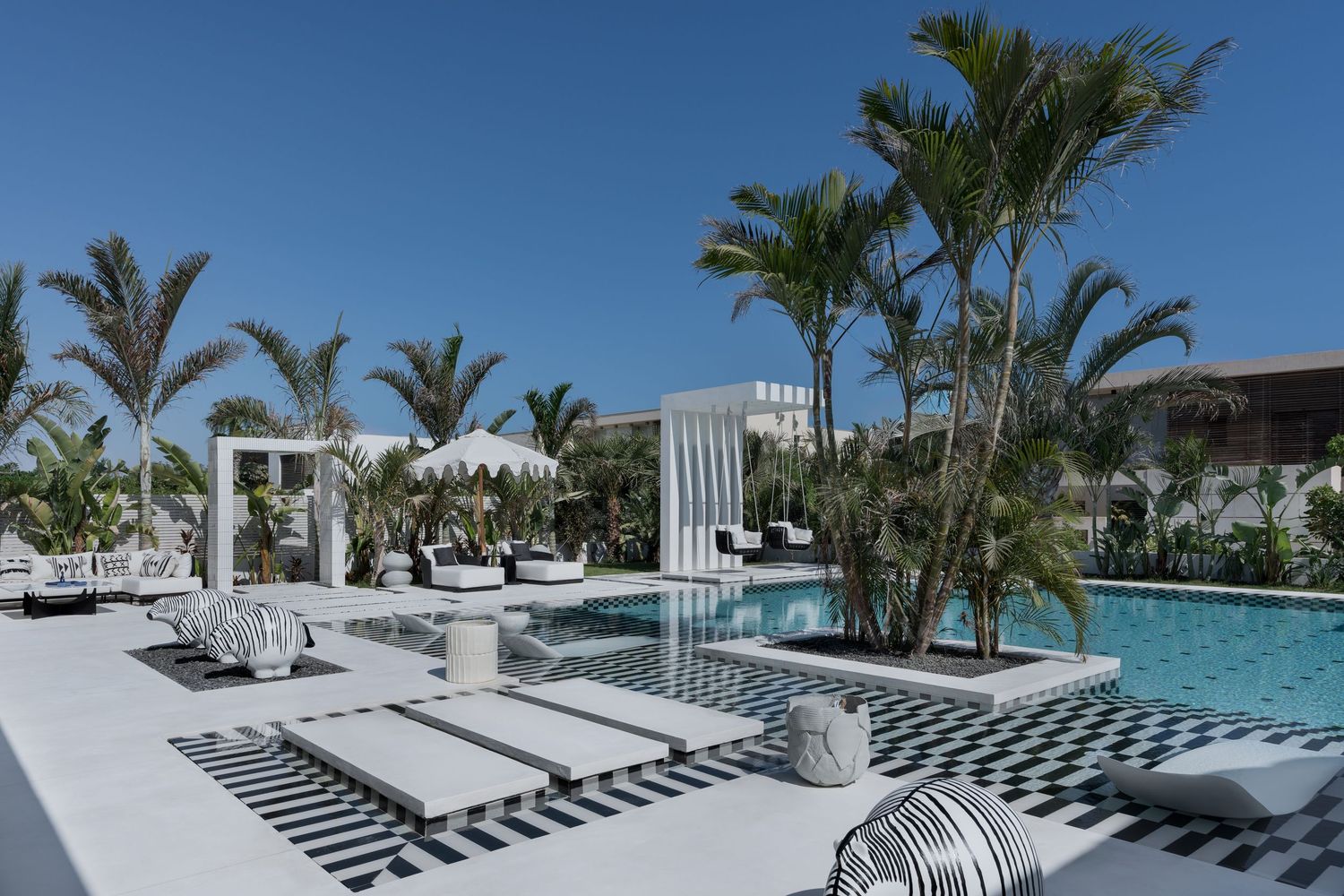 Luxury resort pool area with black and white checkered tiles, white lounge chairs, palm trees, and cabanas under blue sky.