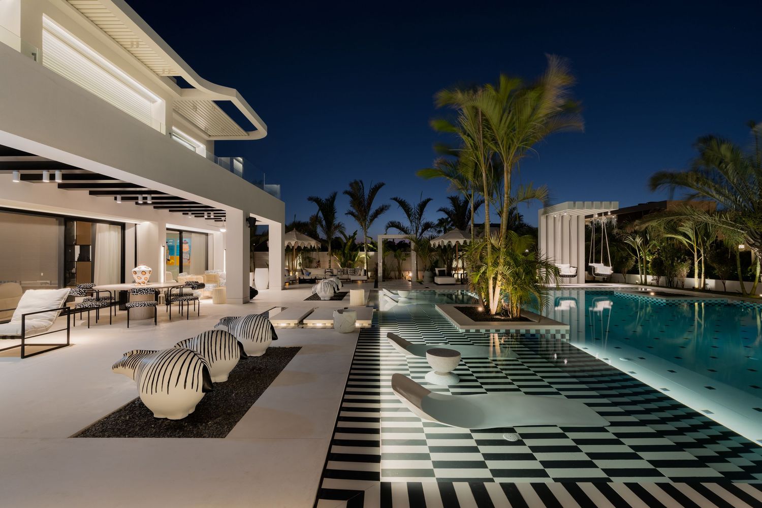 Luxury villa at night with bold black and white checkered pool deck, palm trees, and glowing illuminated swimming pool.
