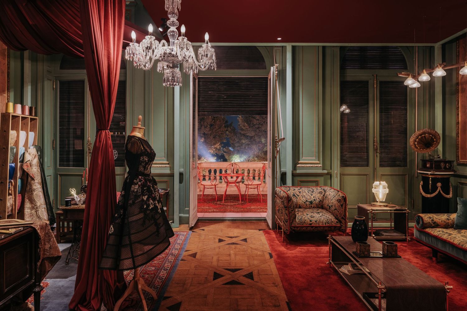 Opulent vintage room with chandelier, green paneled walls, red carpet, mannequin in black gown, and open balcony doorway.