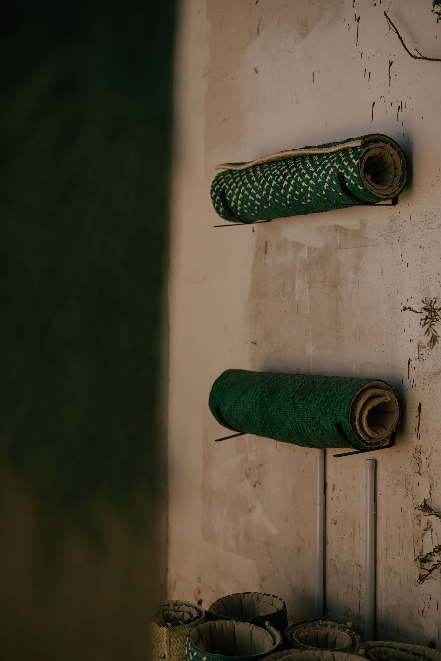 Two rolled green woven mats mounted on wall pegs against a distressed white wall with small baskets below.
