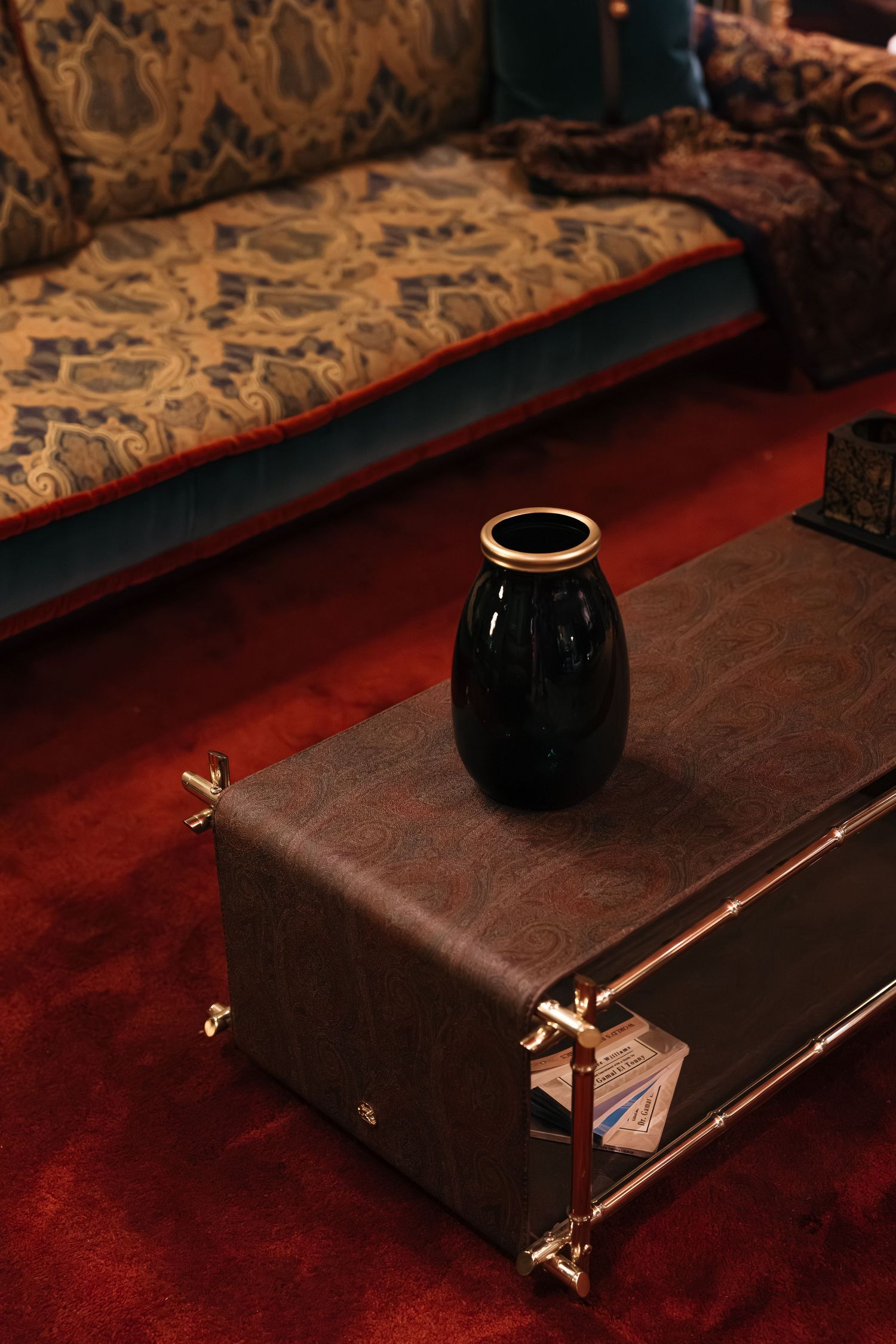 Black ceramic vase on a leather coffee table with ornate patterned sofa on red carpet.