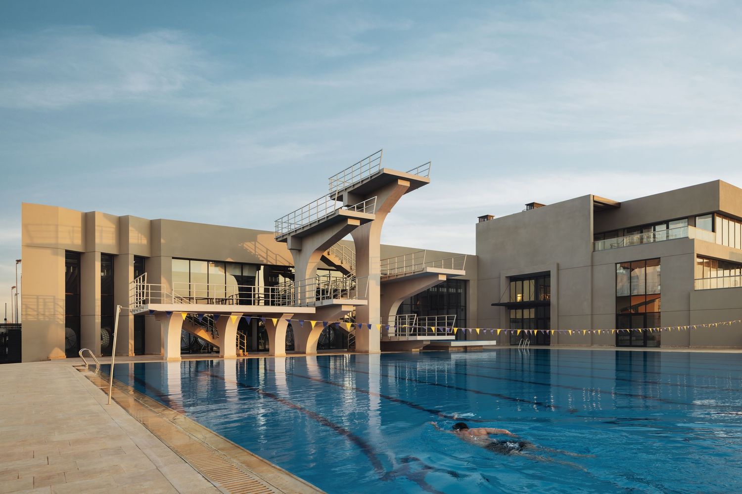 Modern aquatic sports complex with an Olympic swimming pool and high diving platform under a clear blue sky.