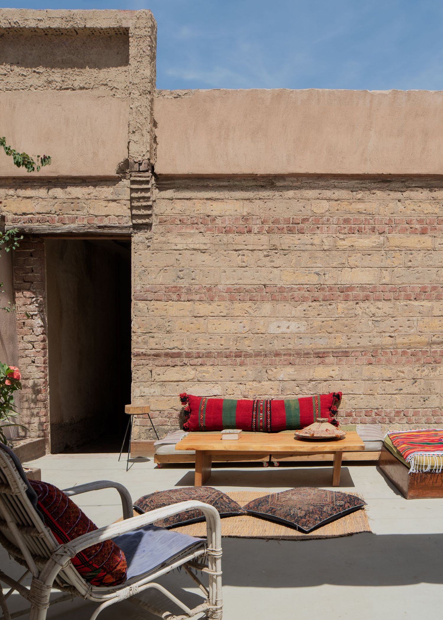 Rustic Moroccan rooftop terrace with colorful kilim cushions, wooden coffee table, and exposed brick walls under blue sky.