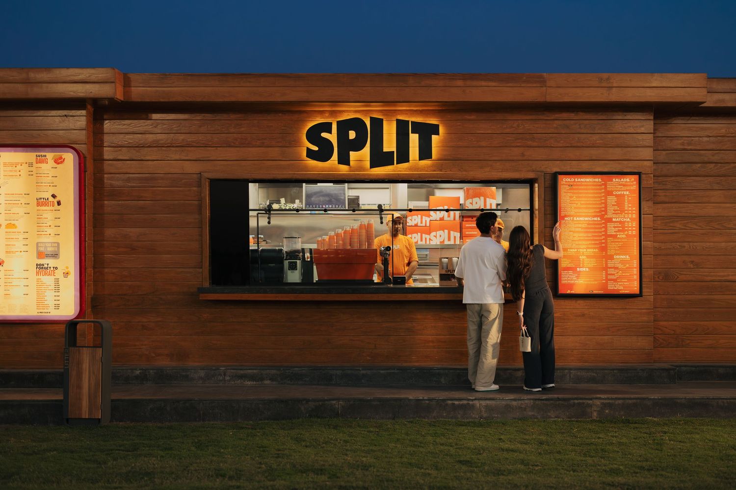 Customers ordering at the illuminated walk-up window of a wooden Split fast food restaurant at dusk.
