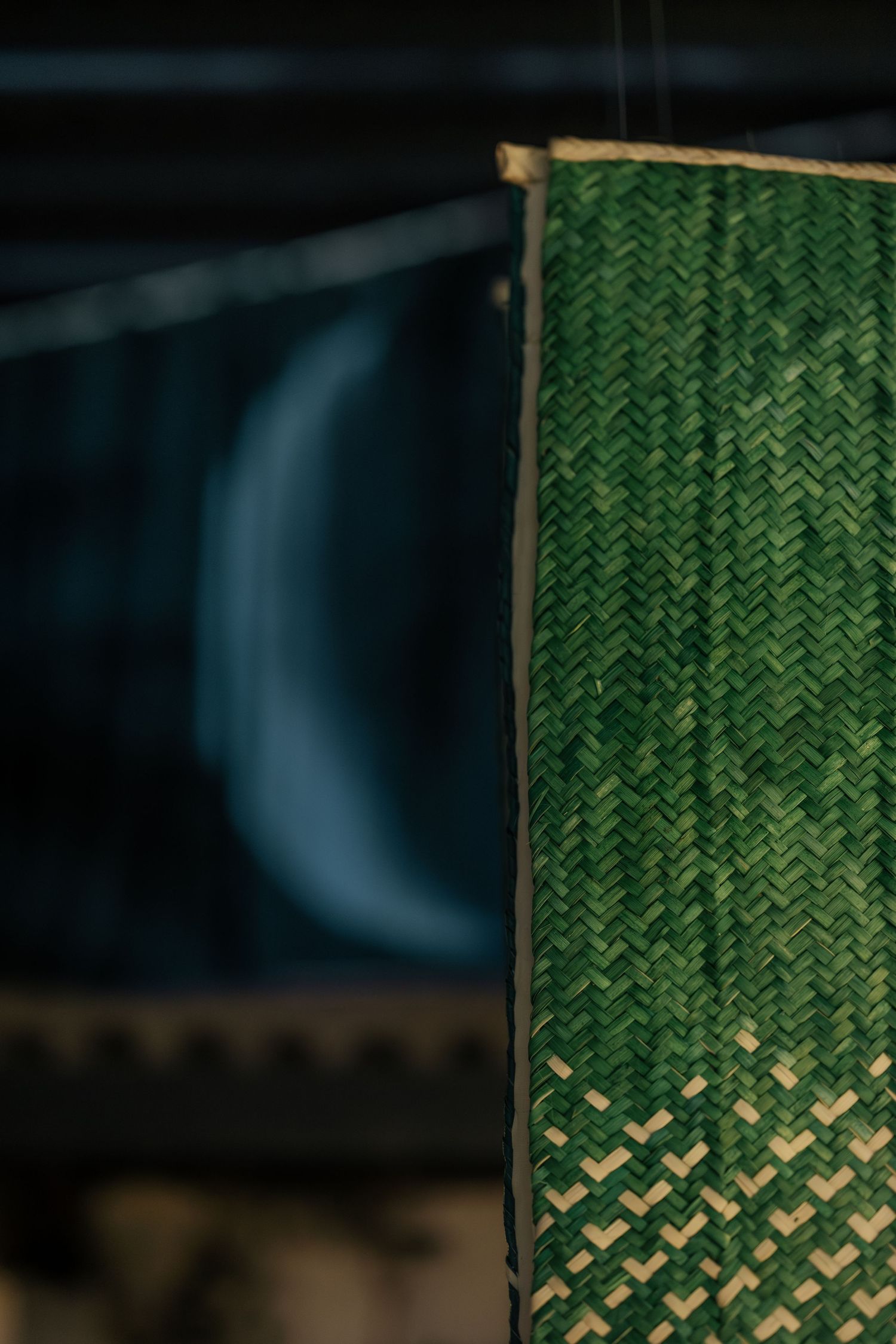 Close-up of a green woven bamboo mat hanging against a dark background with a blurred crescent shape.