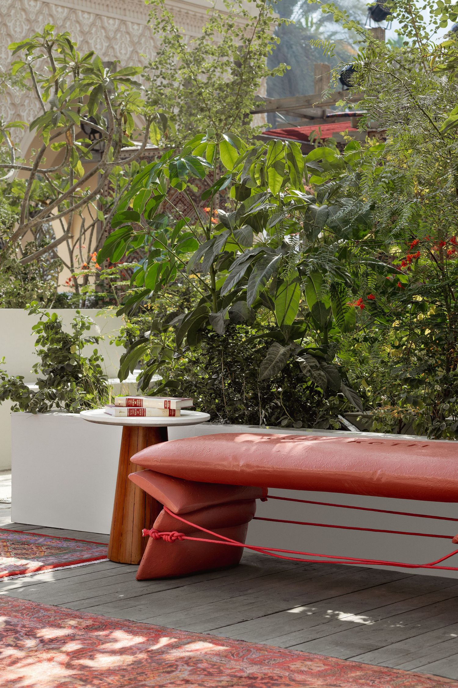 Red cushioned outdoor bench with Persian rug on wooden deck surrounded by lush green tropical plants.