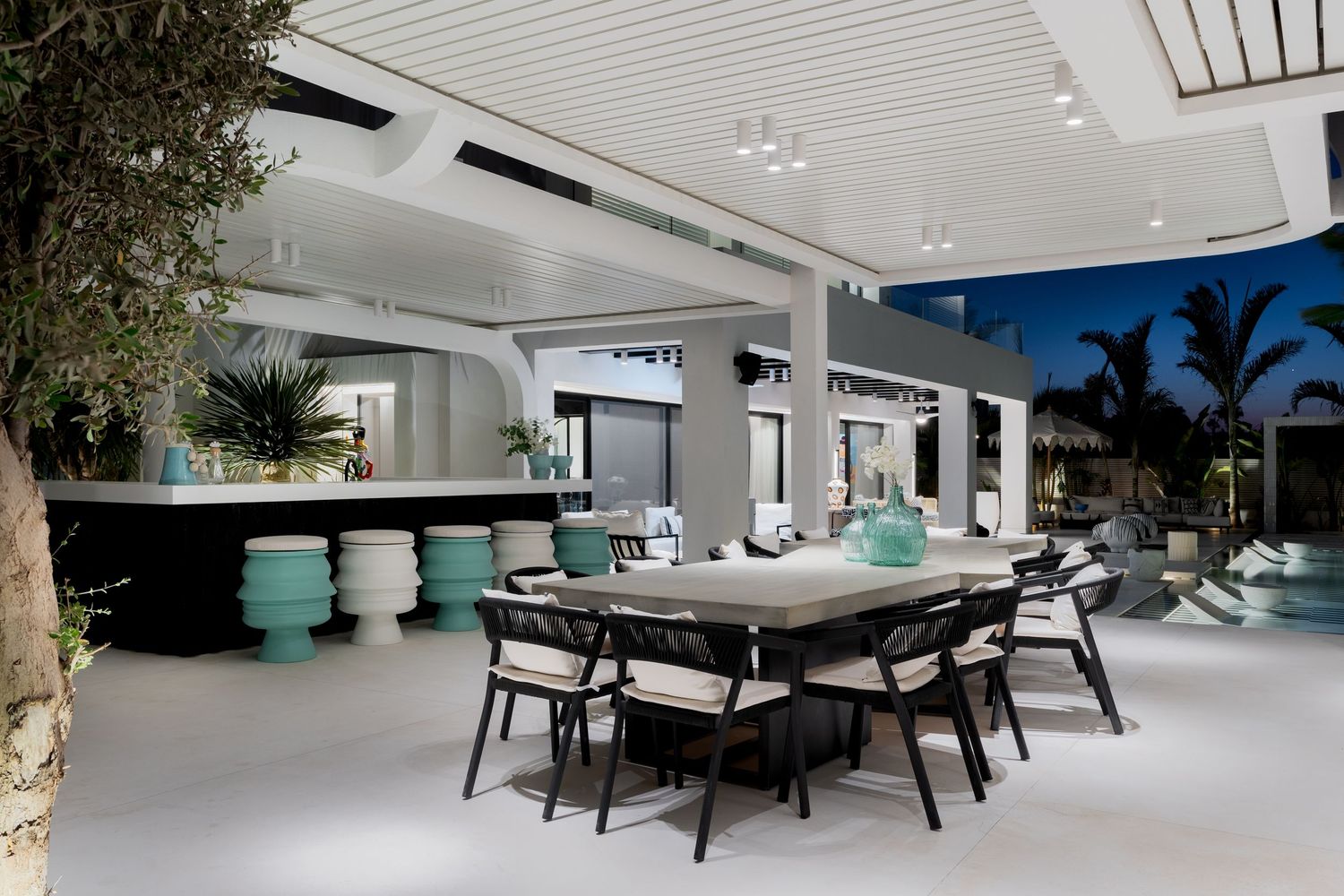 Modern outdoor dining area with black table, teal accent stools, white pergola ceiling, and pool visible in background.