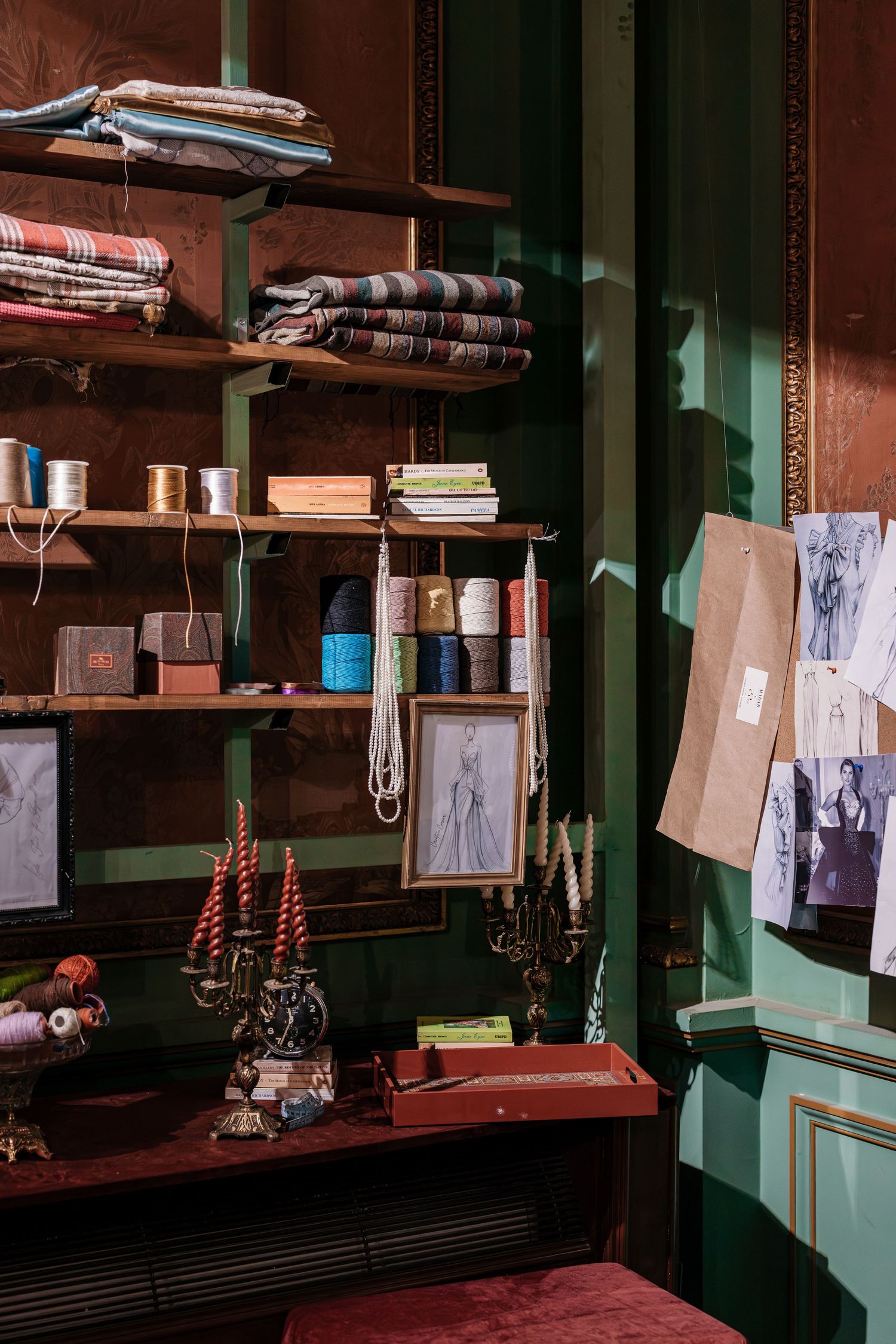 Dark green shelving unit displaying folded fabrics, ribbons, candles and fashion sketches in moody atelier.