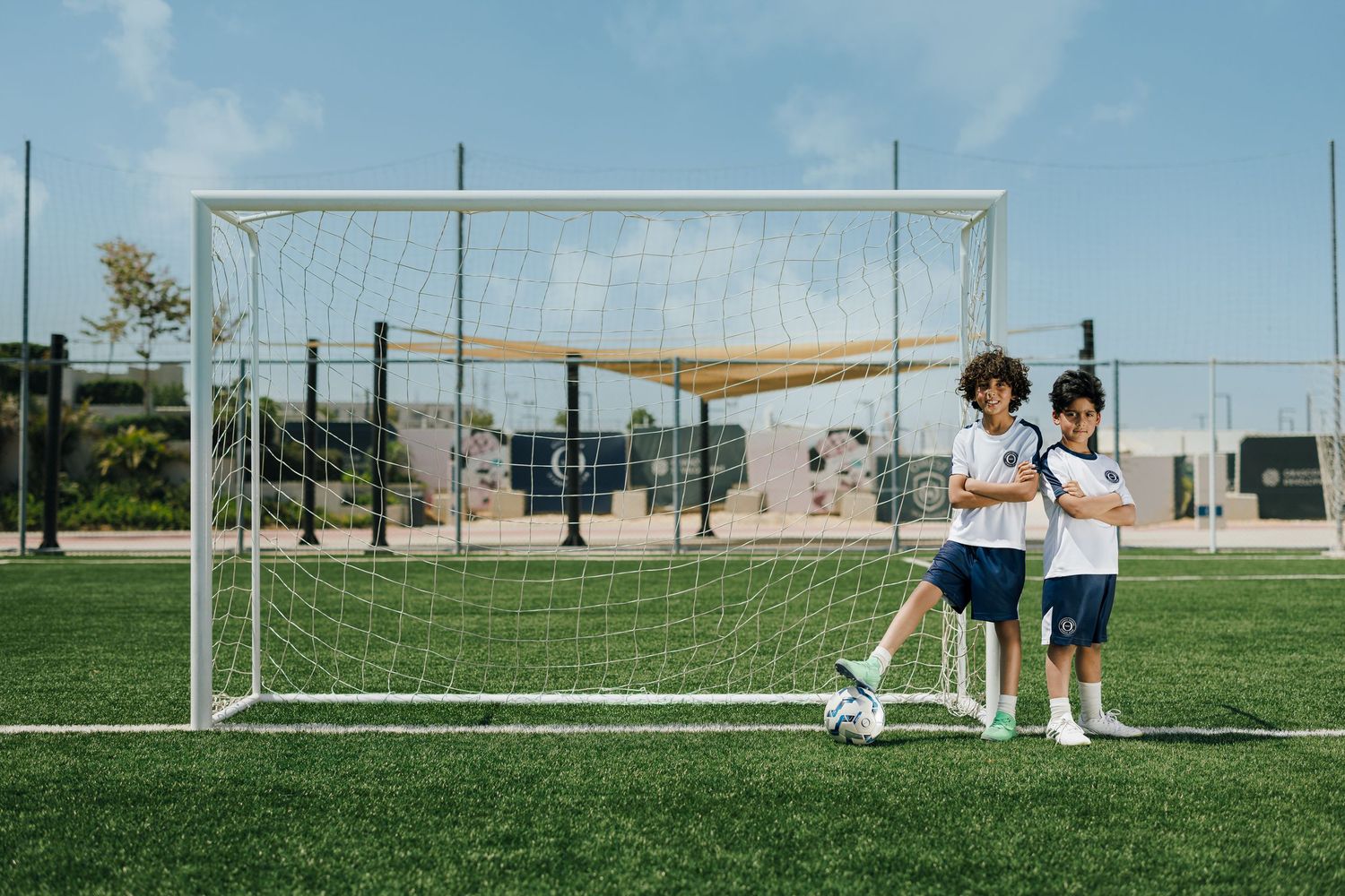 Two young soccer players stand confidently in front of a goal net on a green turf field under a clear blue sky.