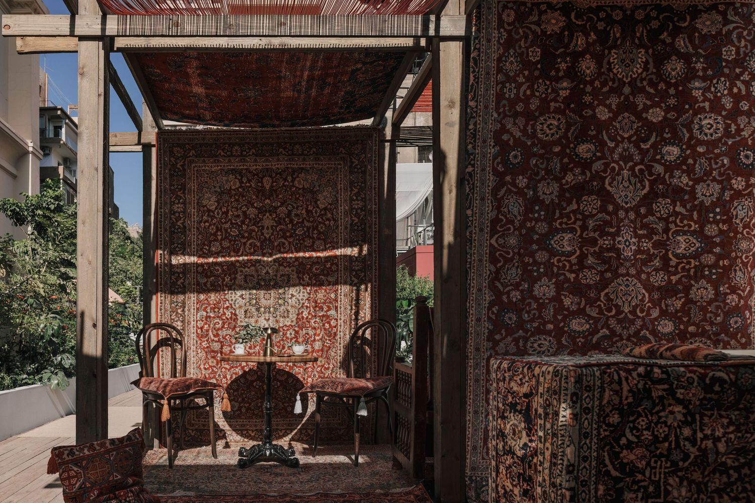 Outdoor pavilion draped with ornate Persian rugs on all sides, enclosing antique wooden chairs in sunlit courtyard.