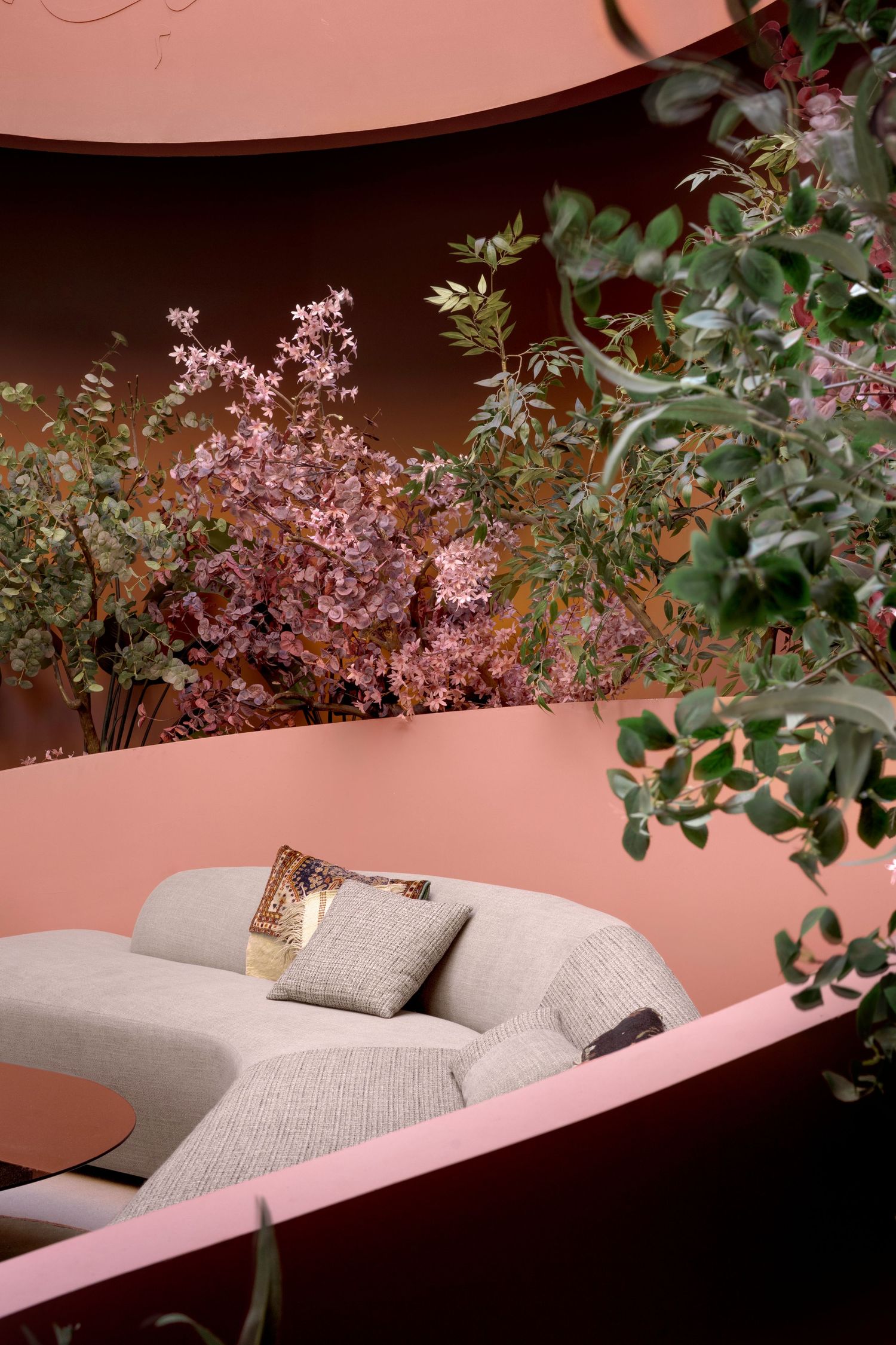 Modern grey sofa with decorative pillows nestled in a pink curved alcove surrounded by lush green and pink plants.