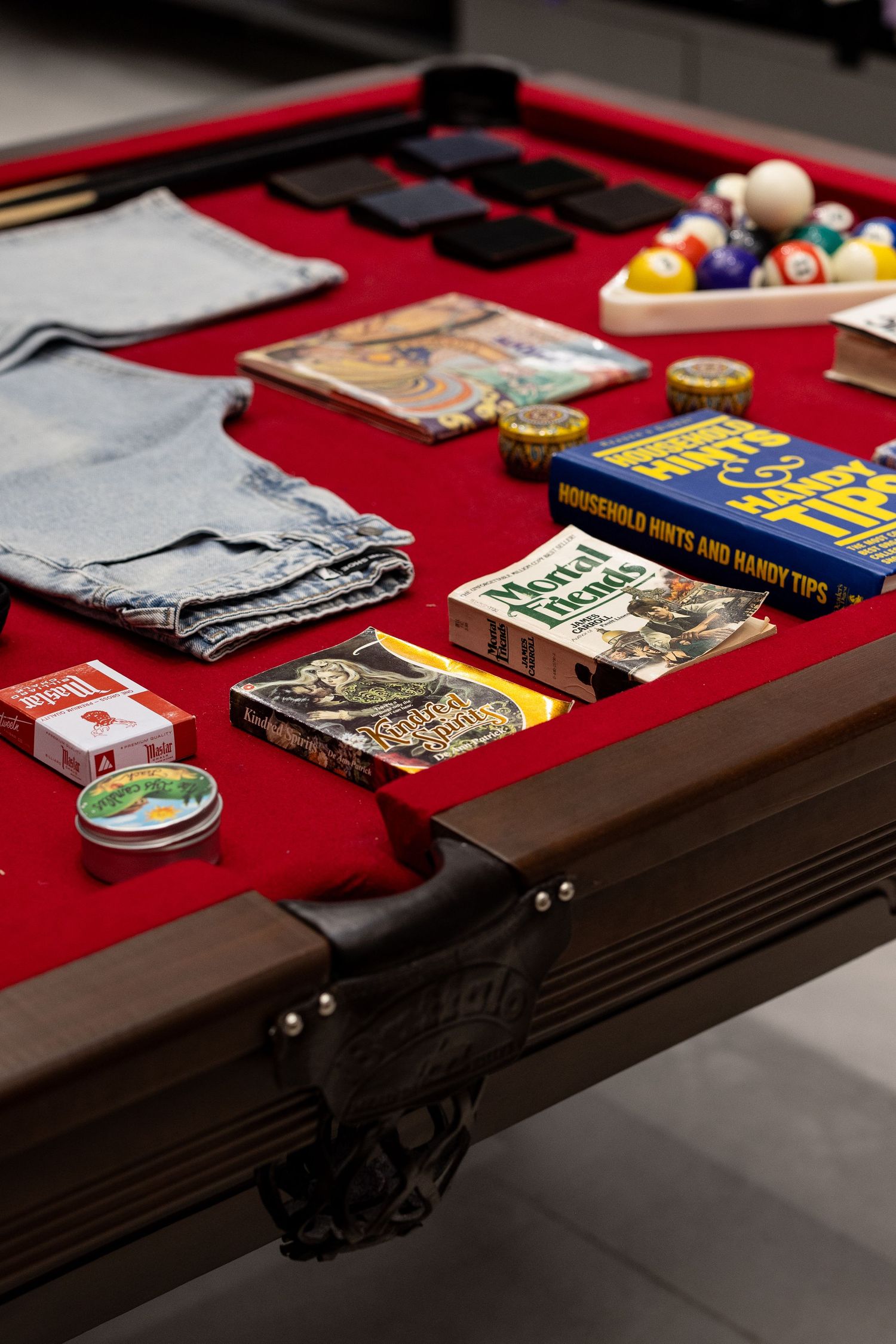 Pool table with red felt displaying jeans, books, billiard balls, and vintage accessories in a retail store.