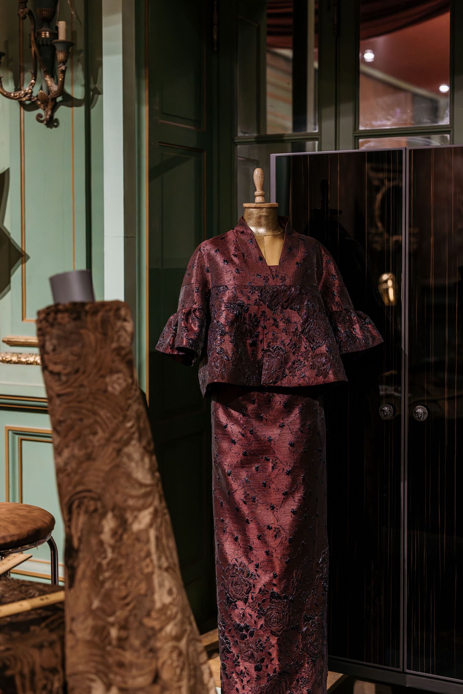 Burgundy brocade two-piece ensemble displayed on a mannequin in an elegant green-paneled room.