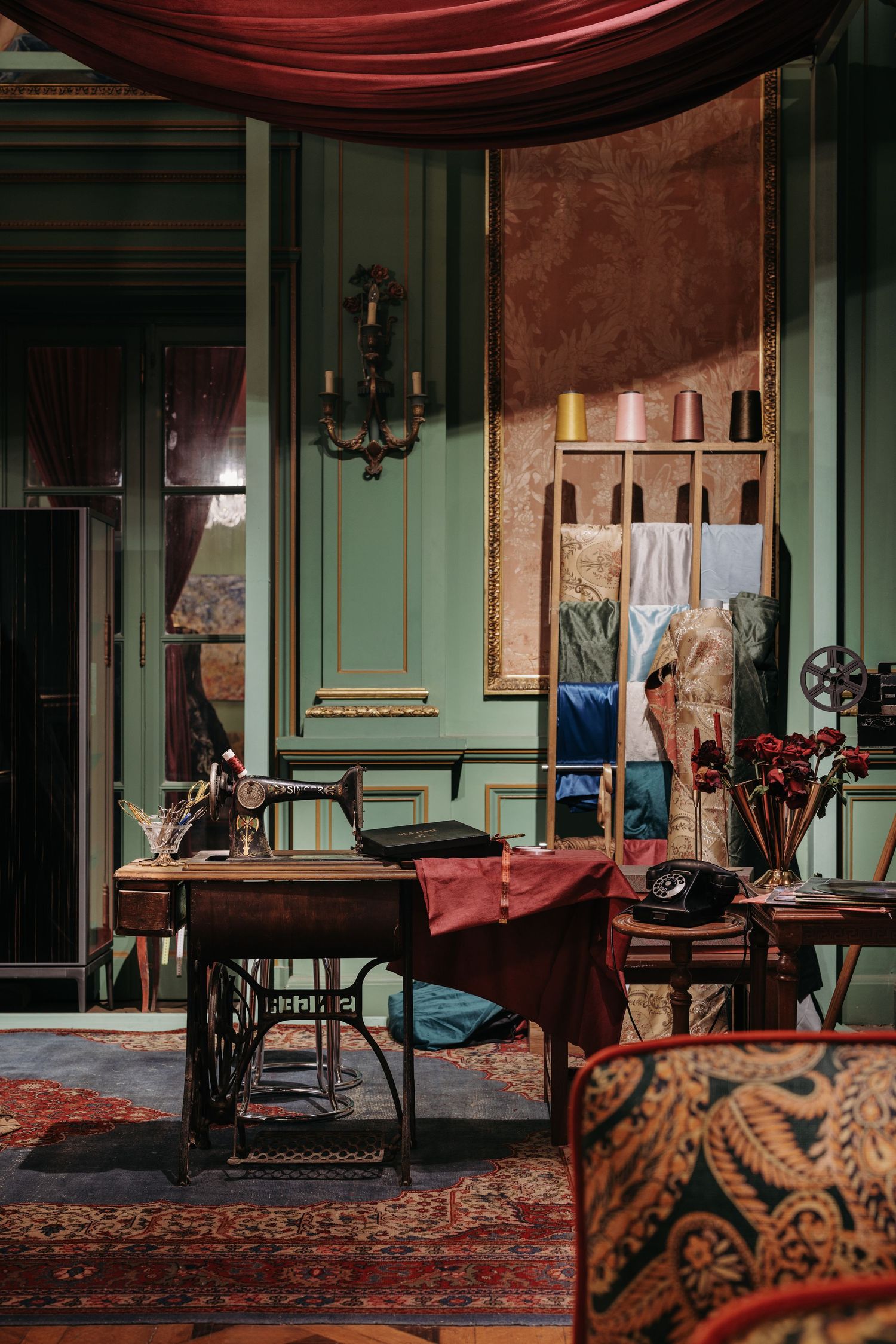 Vintage sewing room with antique treadle machine, red drapes, green walls, and old telephone on ornate carpet.