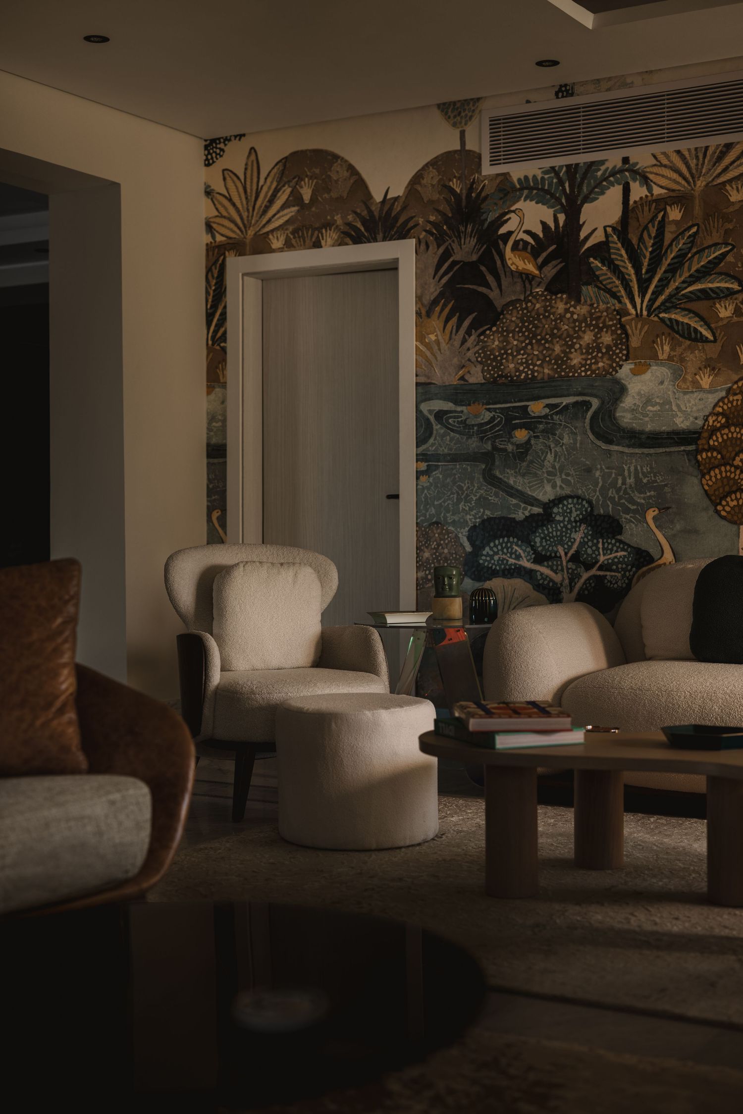 Moody living room with ornate tropical botanical wallpaper mural, cream armchairs, wooden coffee table, and warm lighting.