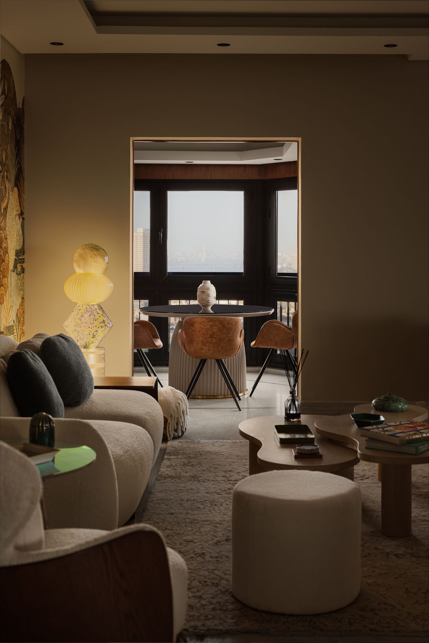 Cozy dimly lit living room with beige sectional sofa, organic coffee table, and tropical mural dining area beyond.