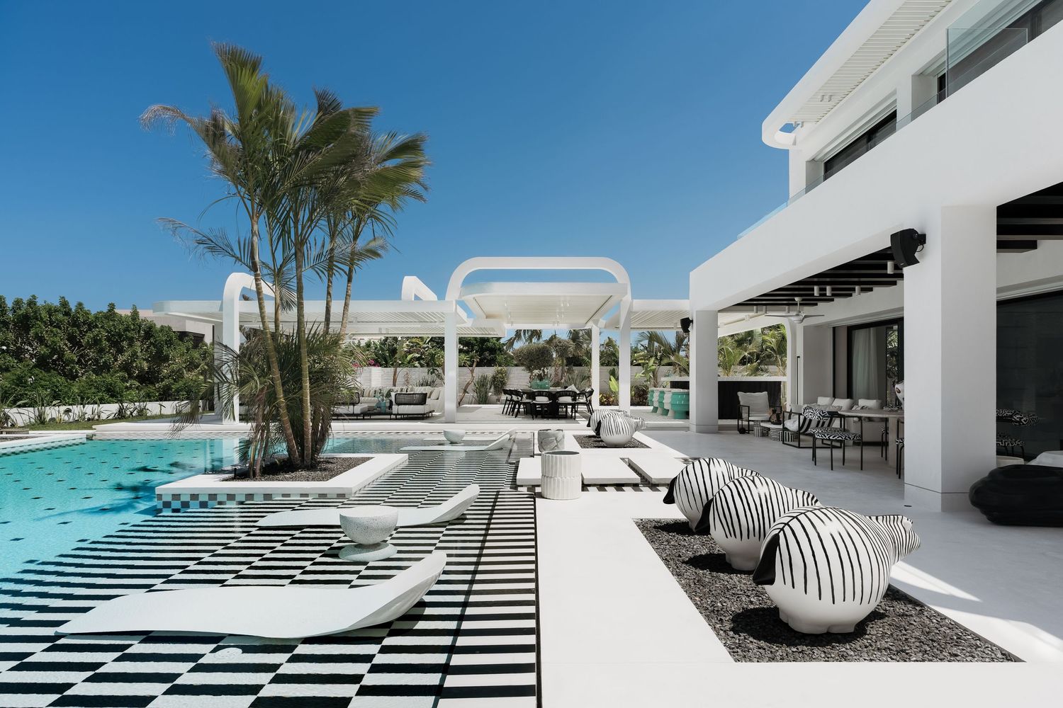 Luxury white villa pool area with striped lounge chairs, palm trees, and modern outdoor furniture under a bright blue sky.