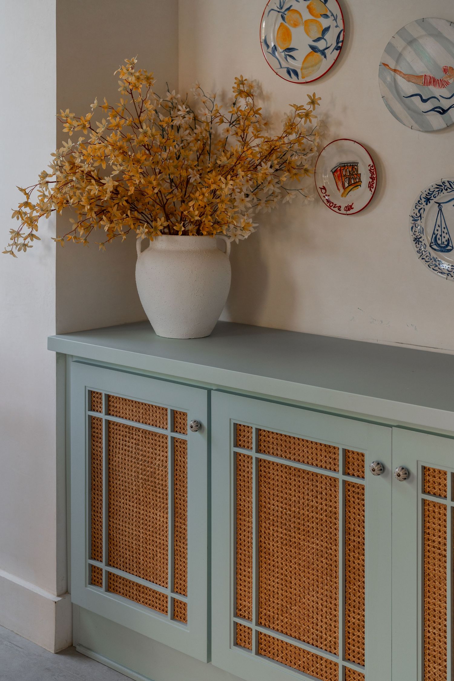 Sage green cabinet with rattan door panels topped with white vase of yellow flowers and decorative wall plates.