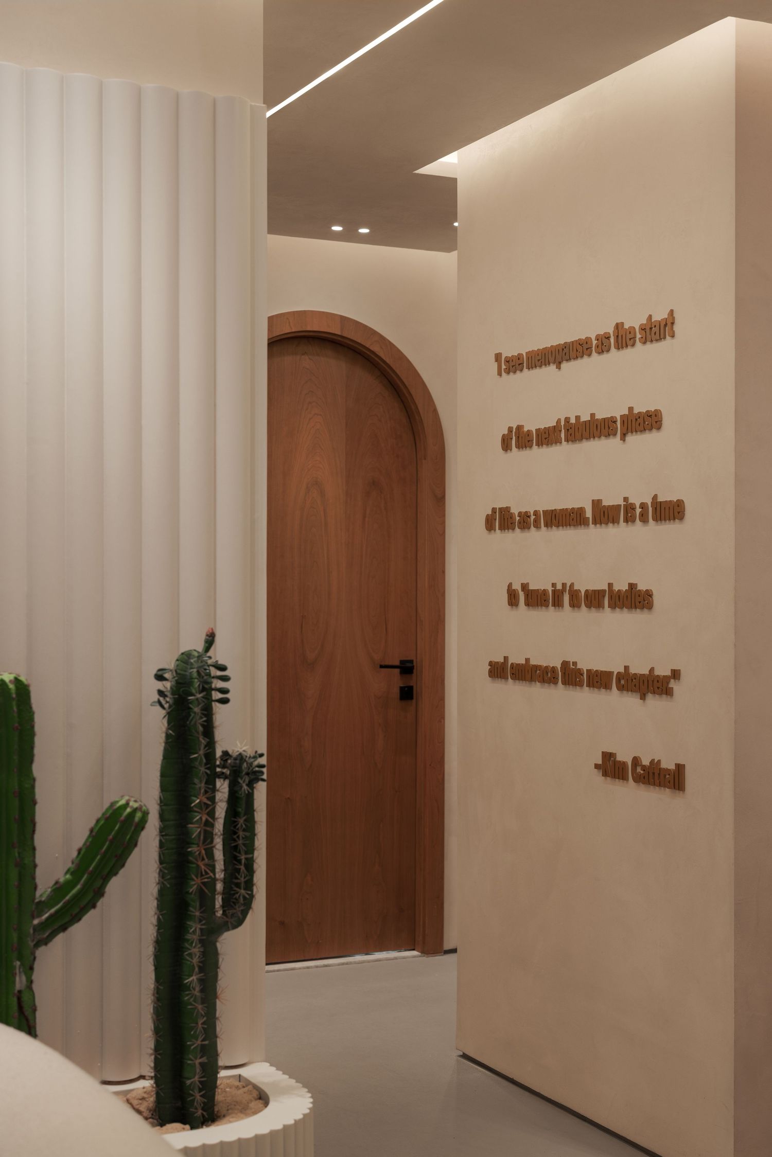 Minimalist corridor with arched wooden door, fluted white panels, cactus plant, and inspirational Kim Cattrall wall quote.