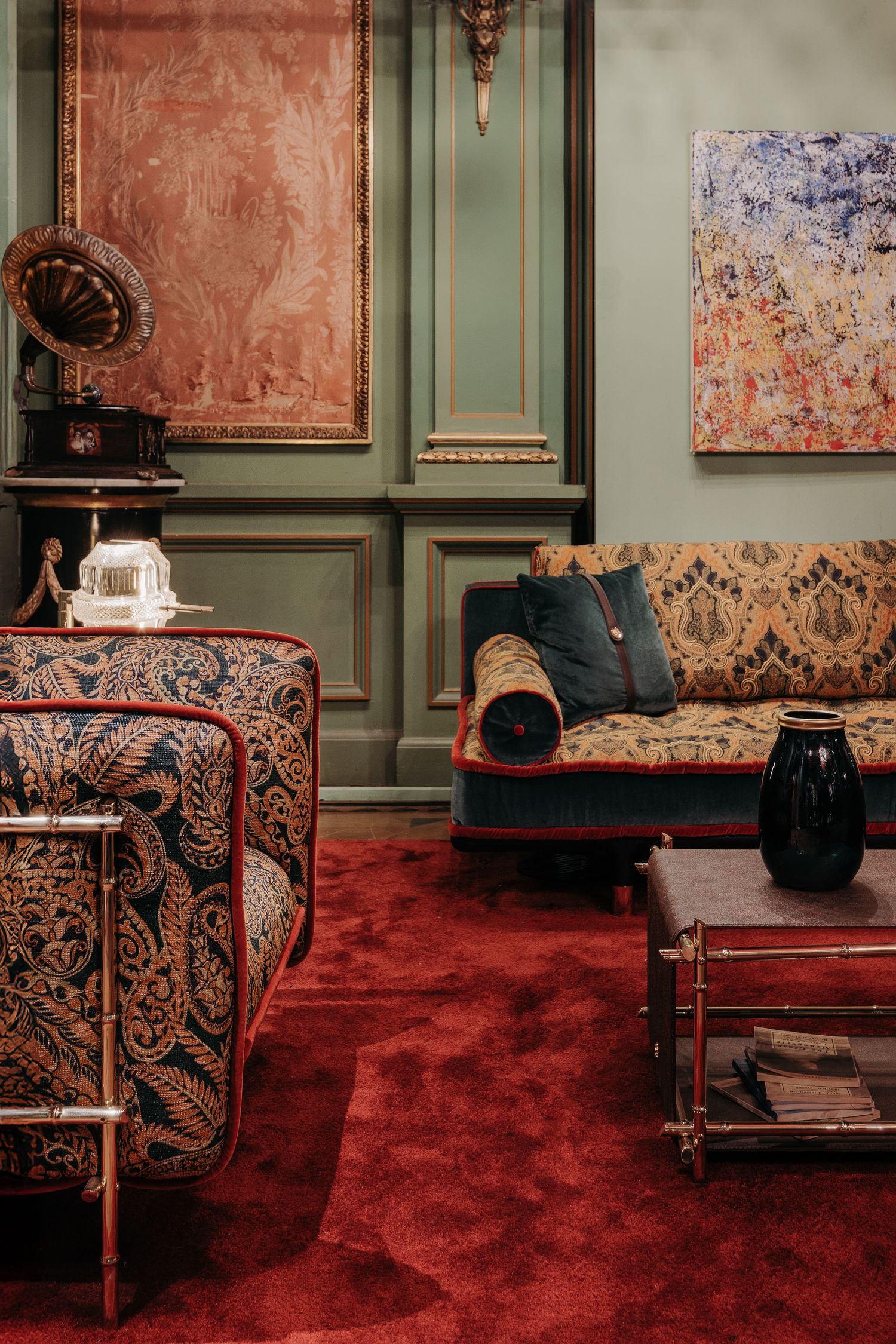 Eclectic vintage living room with ornate patterned armchairs, red carpet, sage green walls, and antique gramophone.