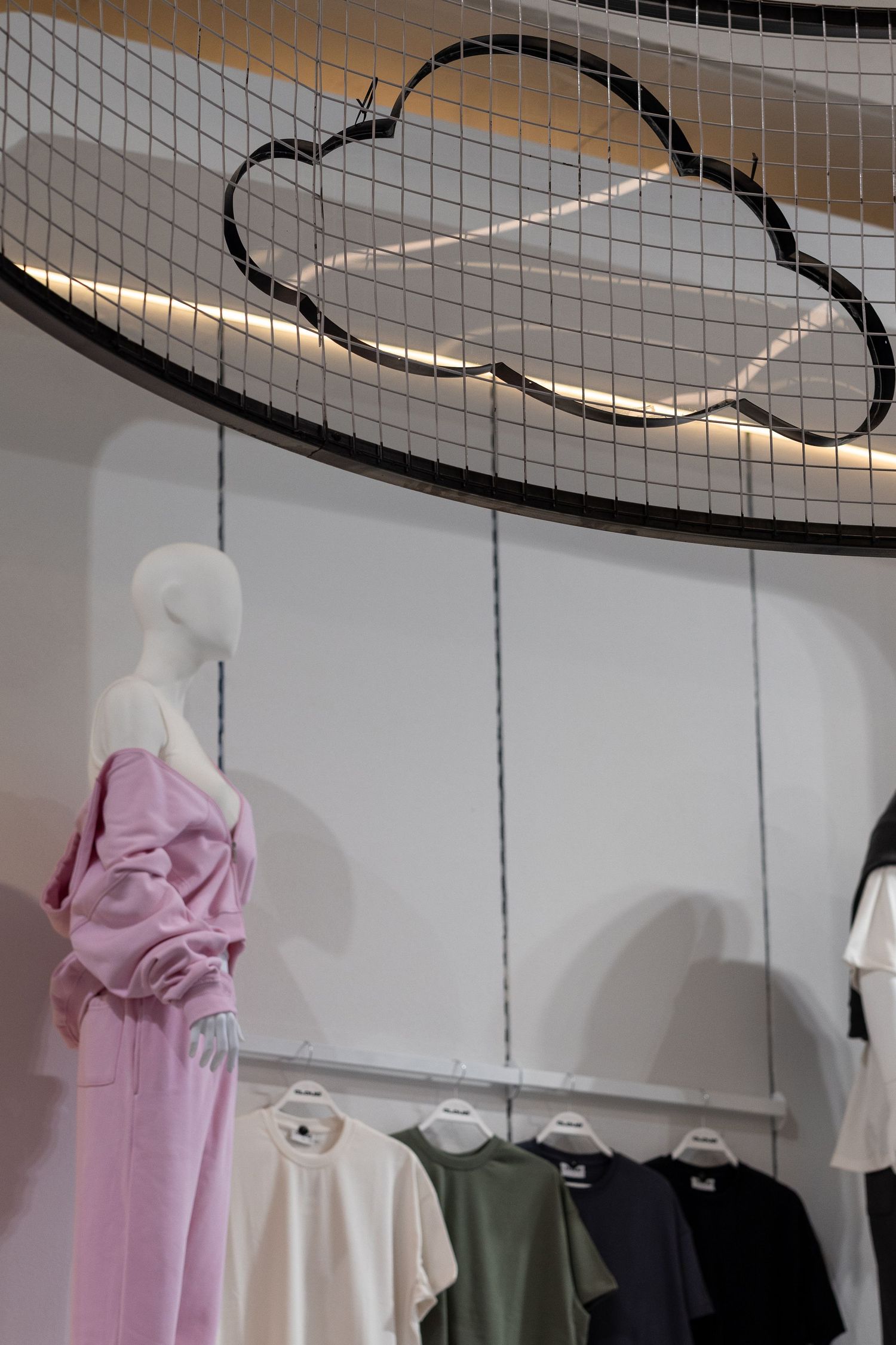 Modern clothing store display with mannequin in pink outfit, hanging t-shirts, and decorative cloud motif ceiling installation.