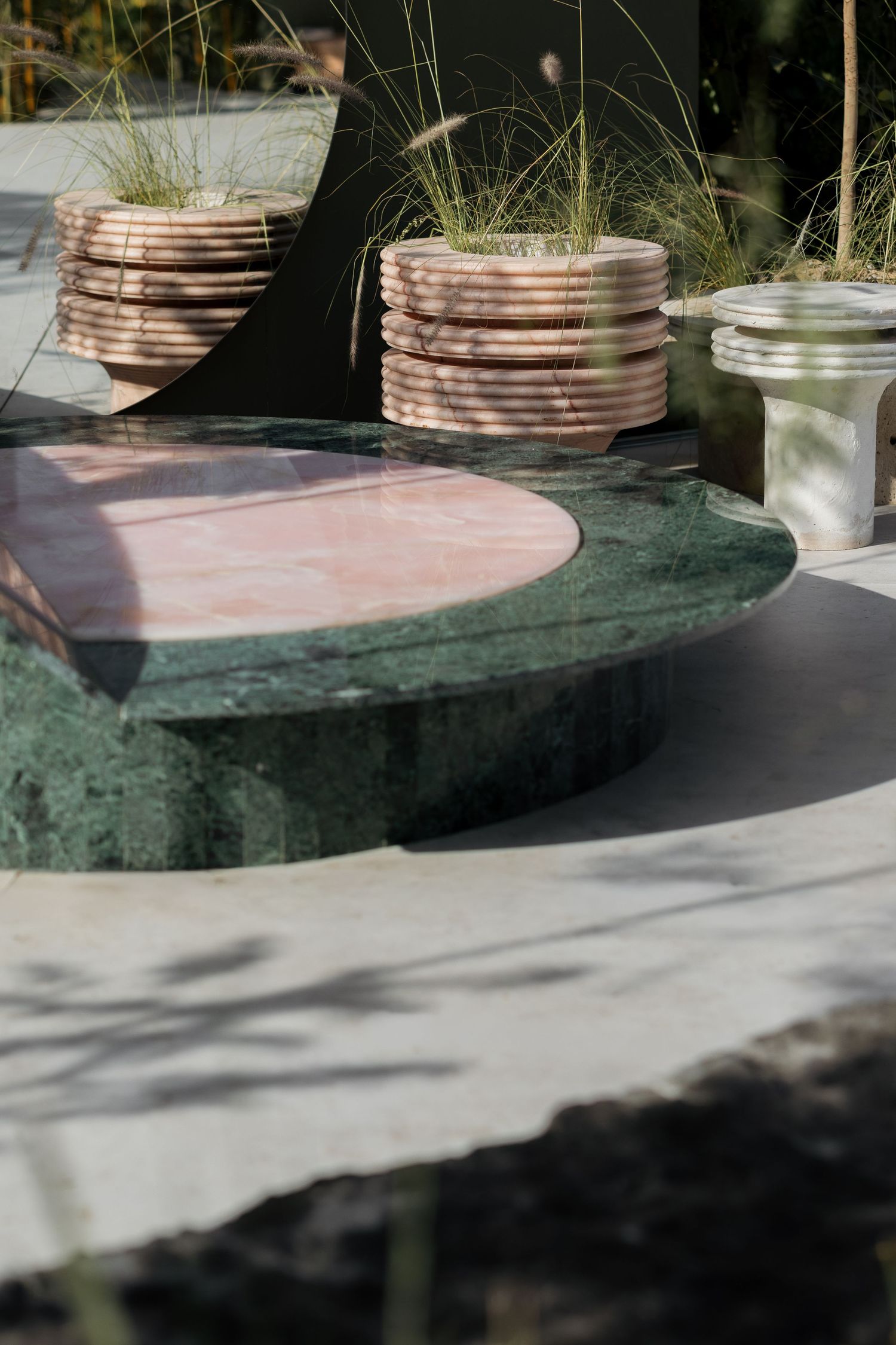 Green and pink marble table with stacked pink stone planters holding ornamental grass in outdoor sculpture garden.