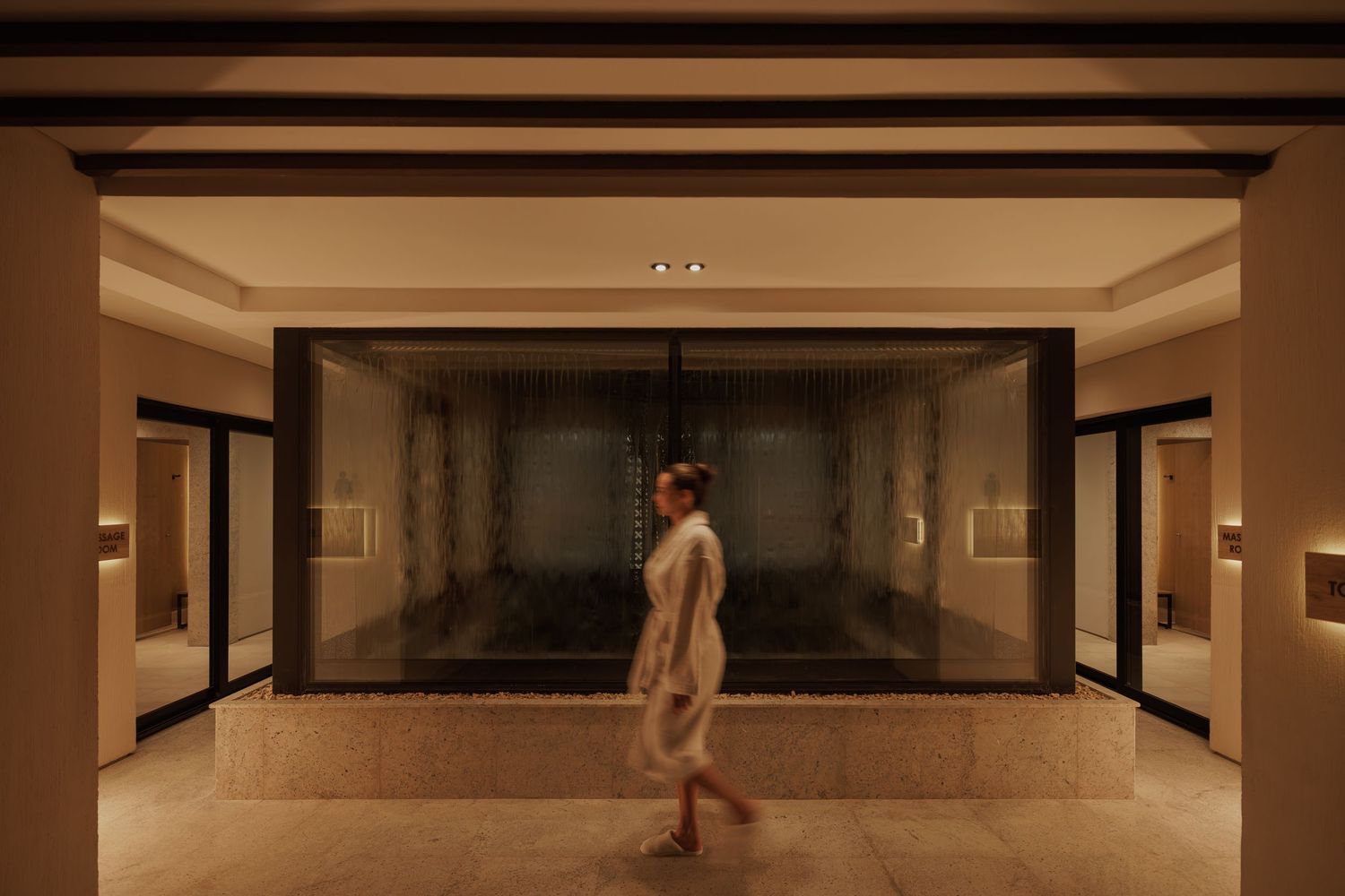 Woman in white robe walking through a dimly lit luxury spa corridor with glass steam room.