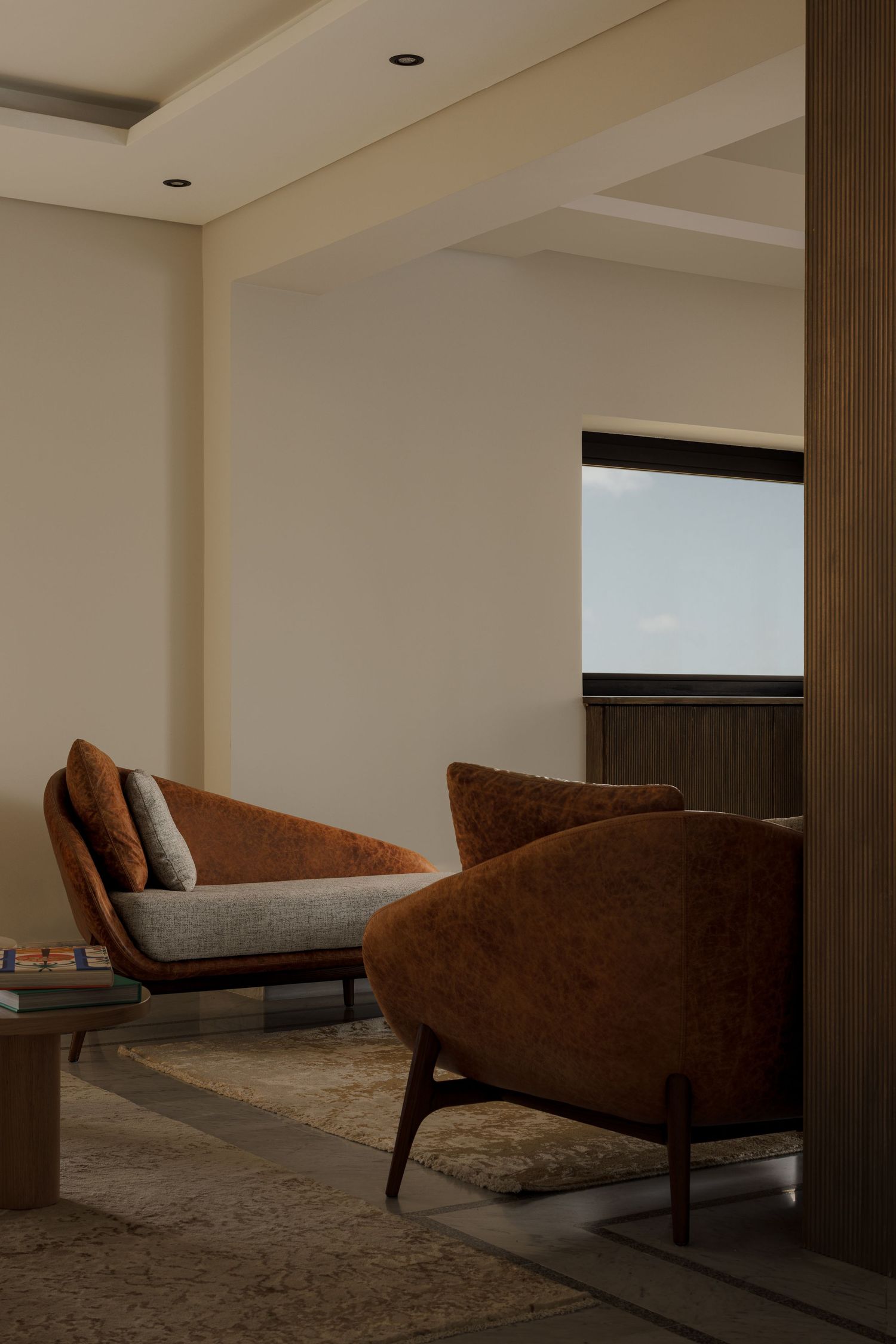 Warm-toned sitting area with rust velvet chairs, beige cushions, recessed lighting, and city view through window.