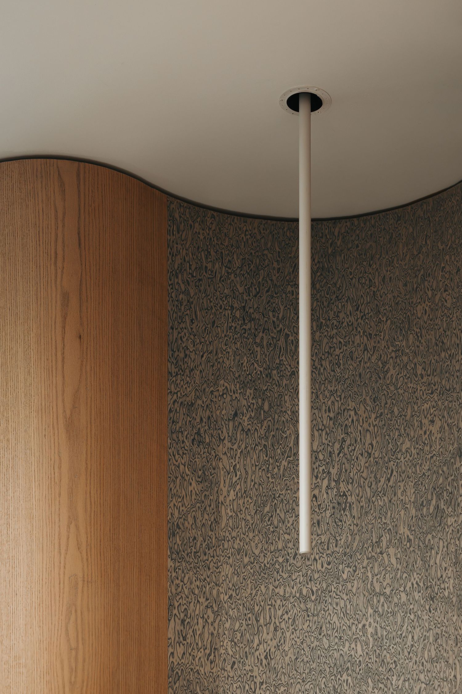 Minimalist interior detail showing curved wood paneling beside textured stone wall with slim pendant light fixture.