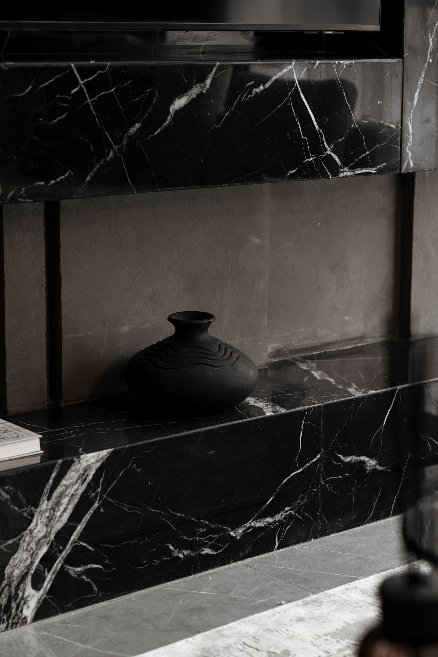 Black matte ceramic vase displayed on a black marble shelf with white veining in a modern interior.