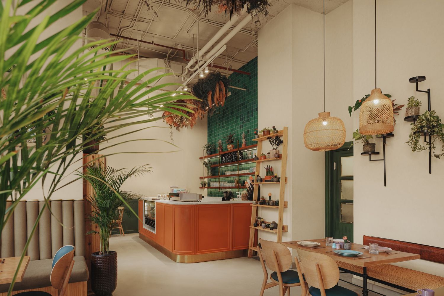 Bright cafe interior with terracotta counter, rattan pendant lights, lush tropical plants, and wooden furniture.