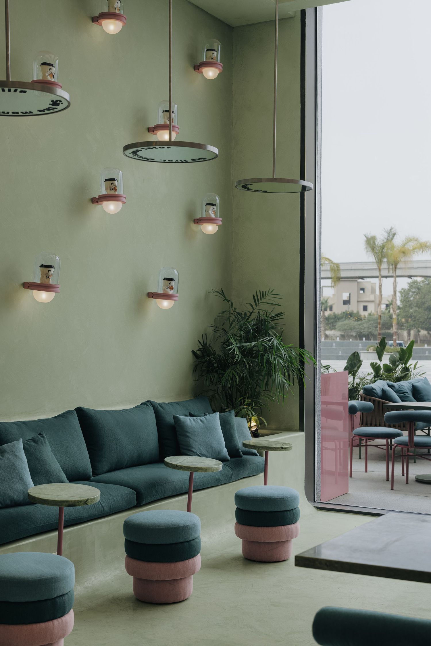 Modern lounge with sage green walls, teal sofas, pink tiered ottomans, and unique pendant lights by large windows.