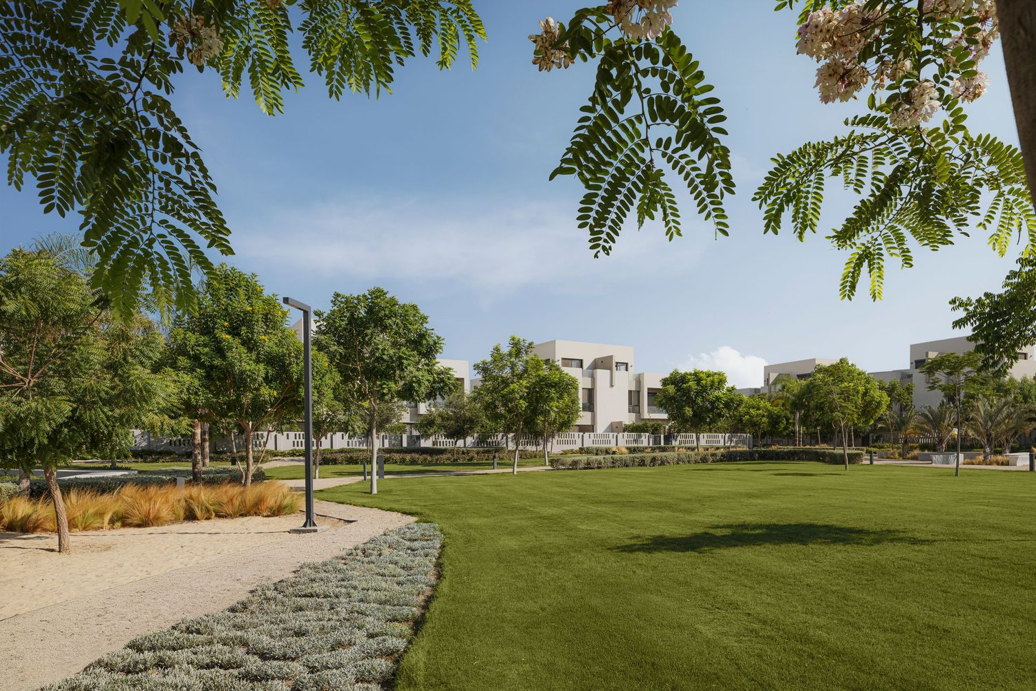 Expansive green park with manicured lawn, trees, and stone-edged pathways near modern buildings under a sunny sky.