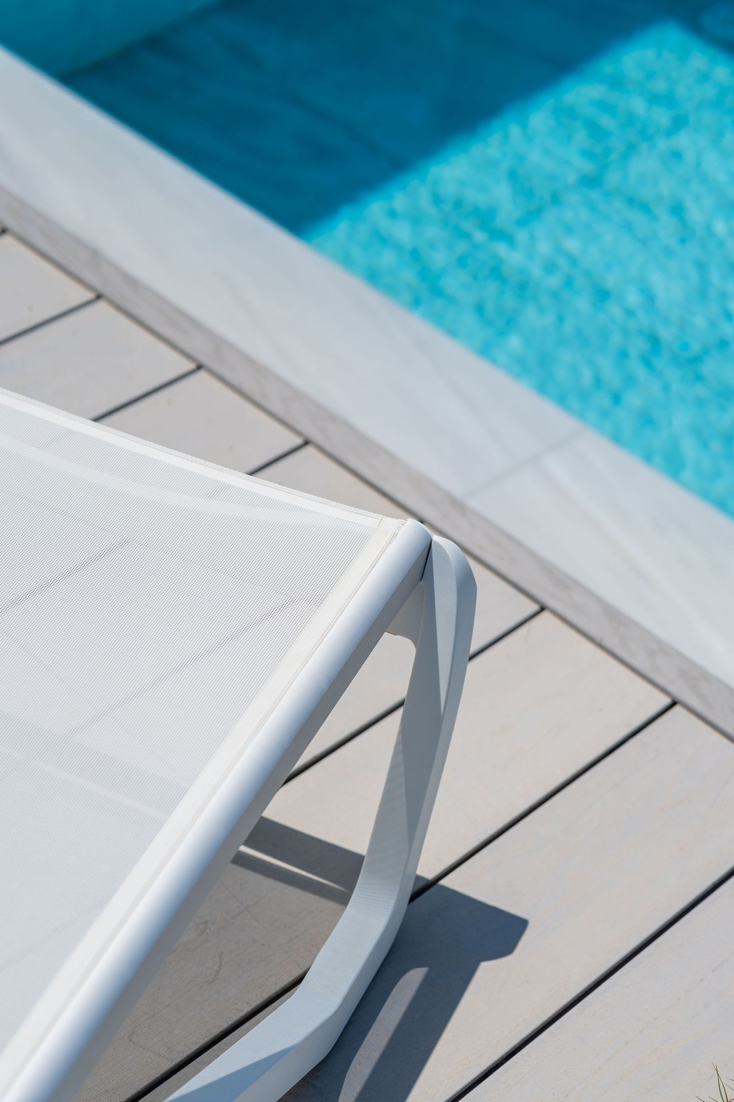 White lounge chair on gray wood deck beside a sparkling blue swimming pool in bright sunlight.