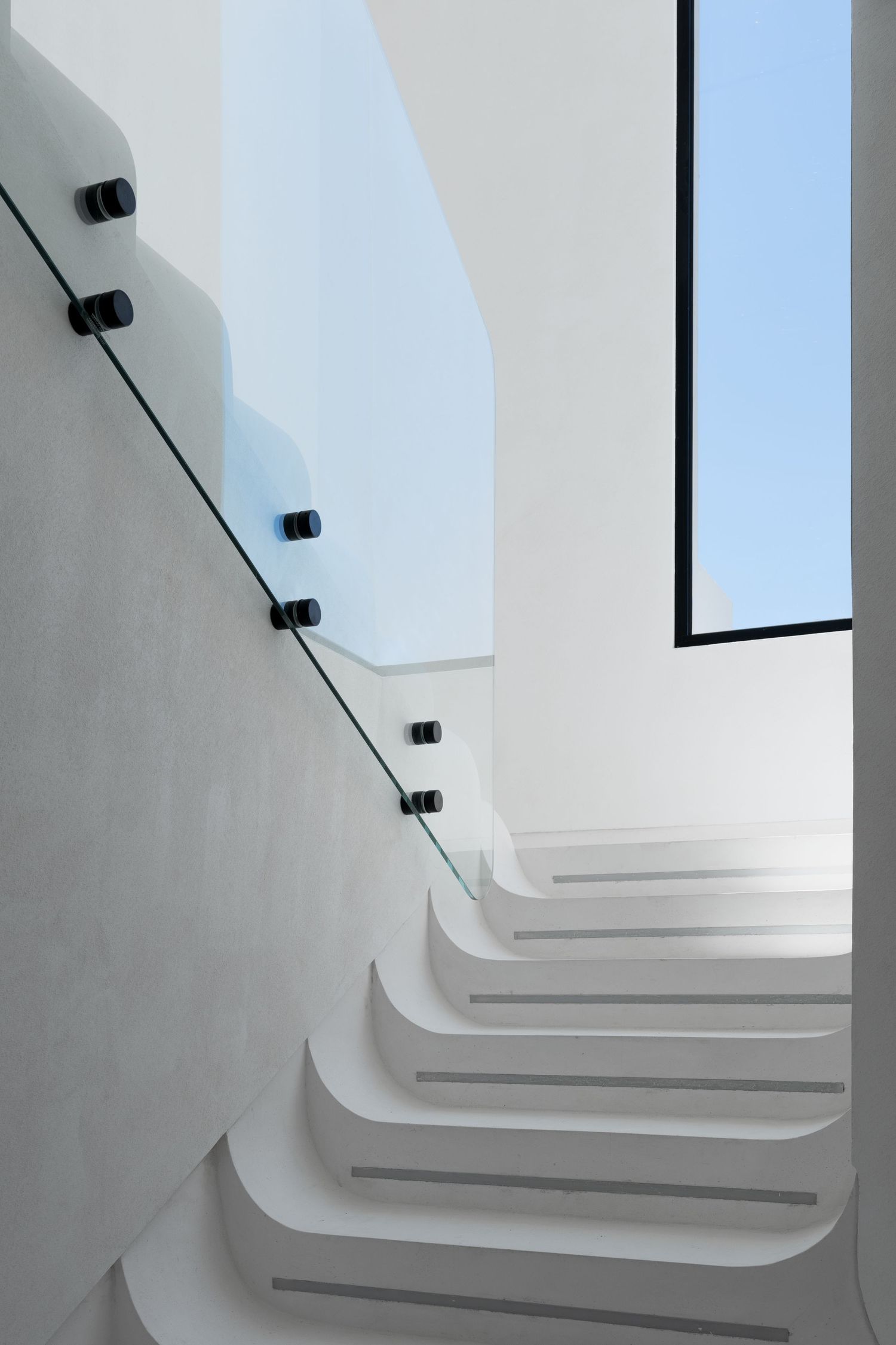 Sleek white curved staircase with glass and black metal railing leading toward a bright skylight window.