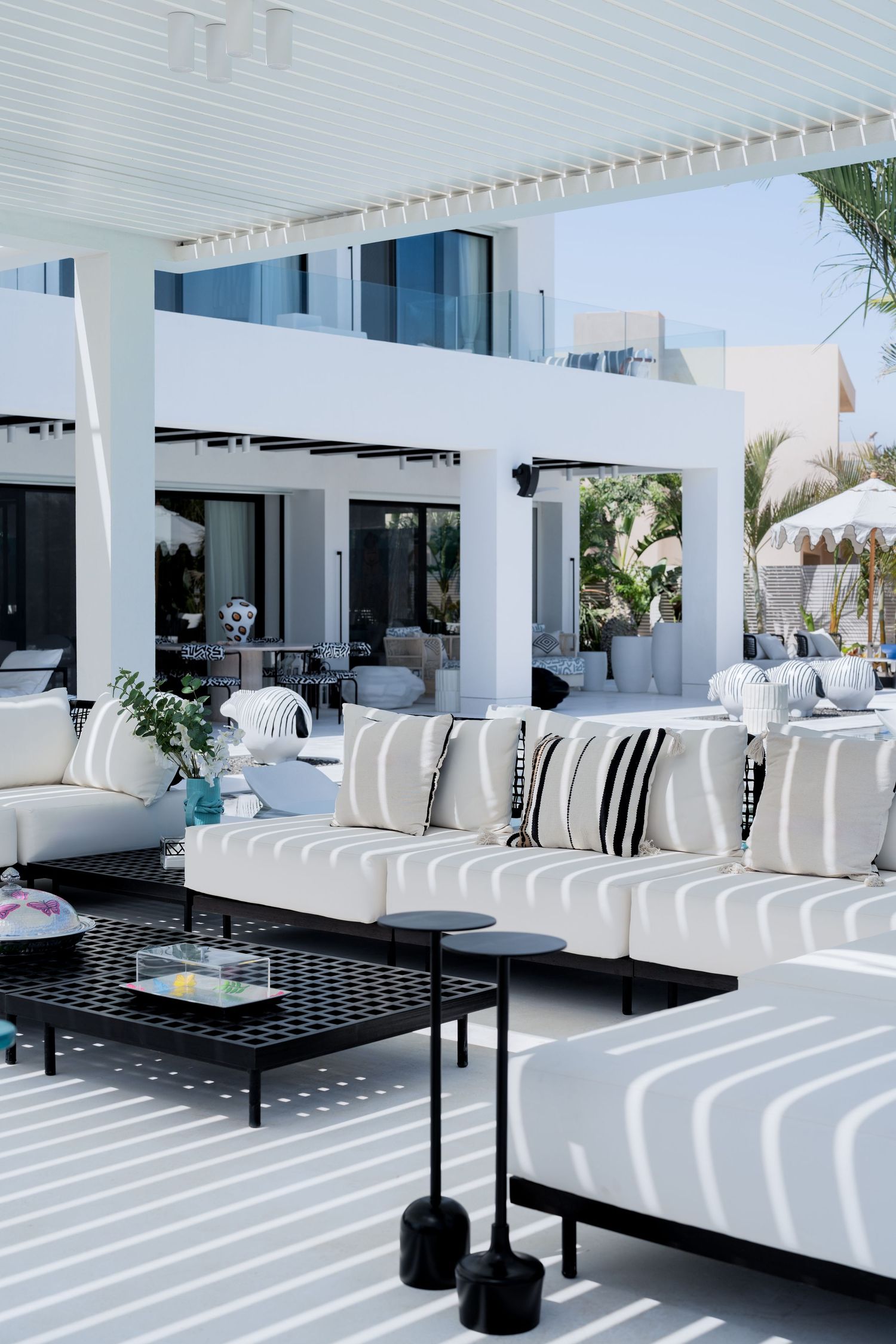 Elegant white outdoor sofa seating with black coffee tables under a white pergola with striped shadow patterns.