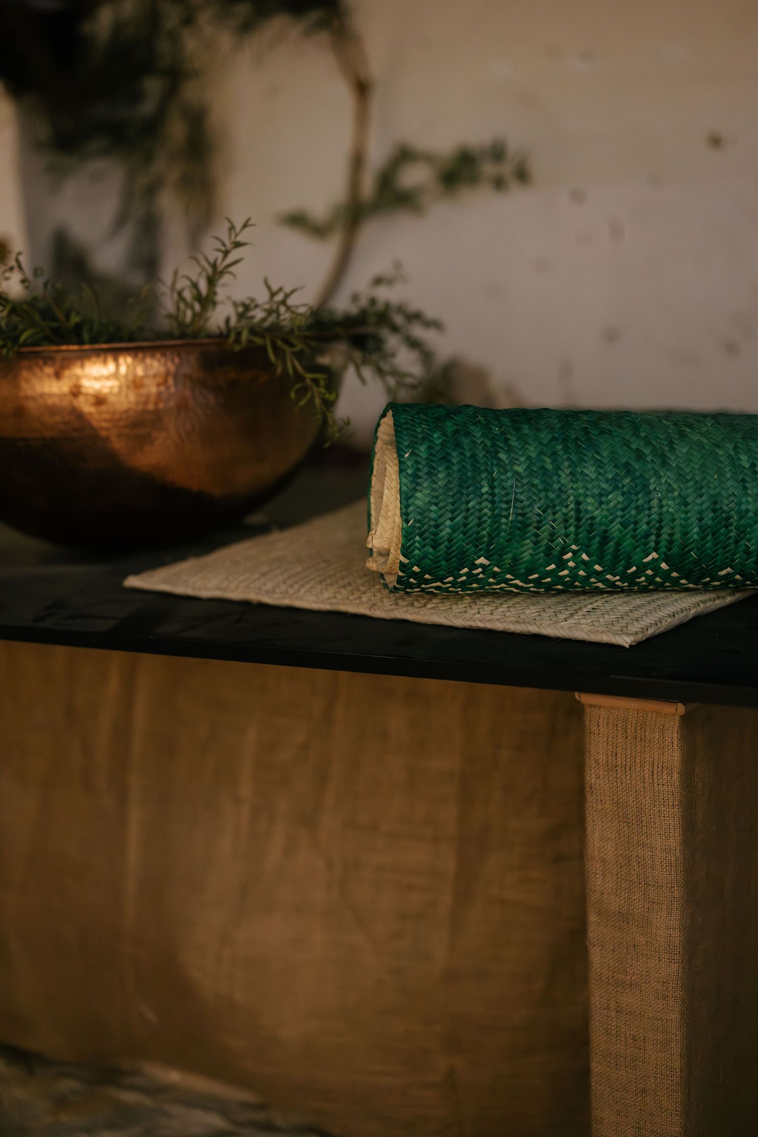 Green woven textile roll resting on burlap-covered surface beside copper bowl with greenery in dim light.