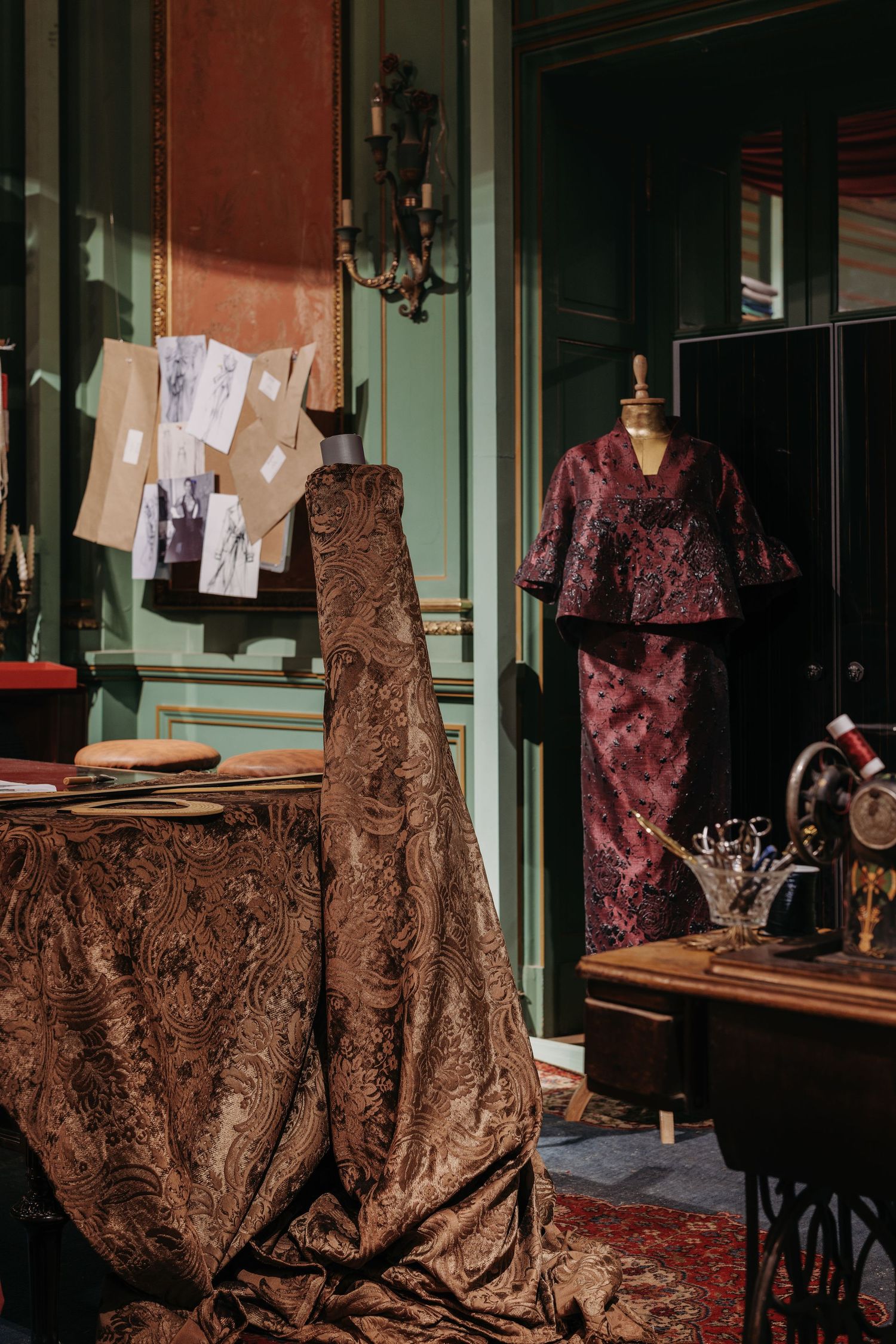Elegant fashion atelier with bronze brocade fabric, mannequin in burgundy dress, and vintage sewing sketches.