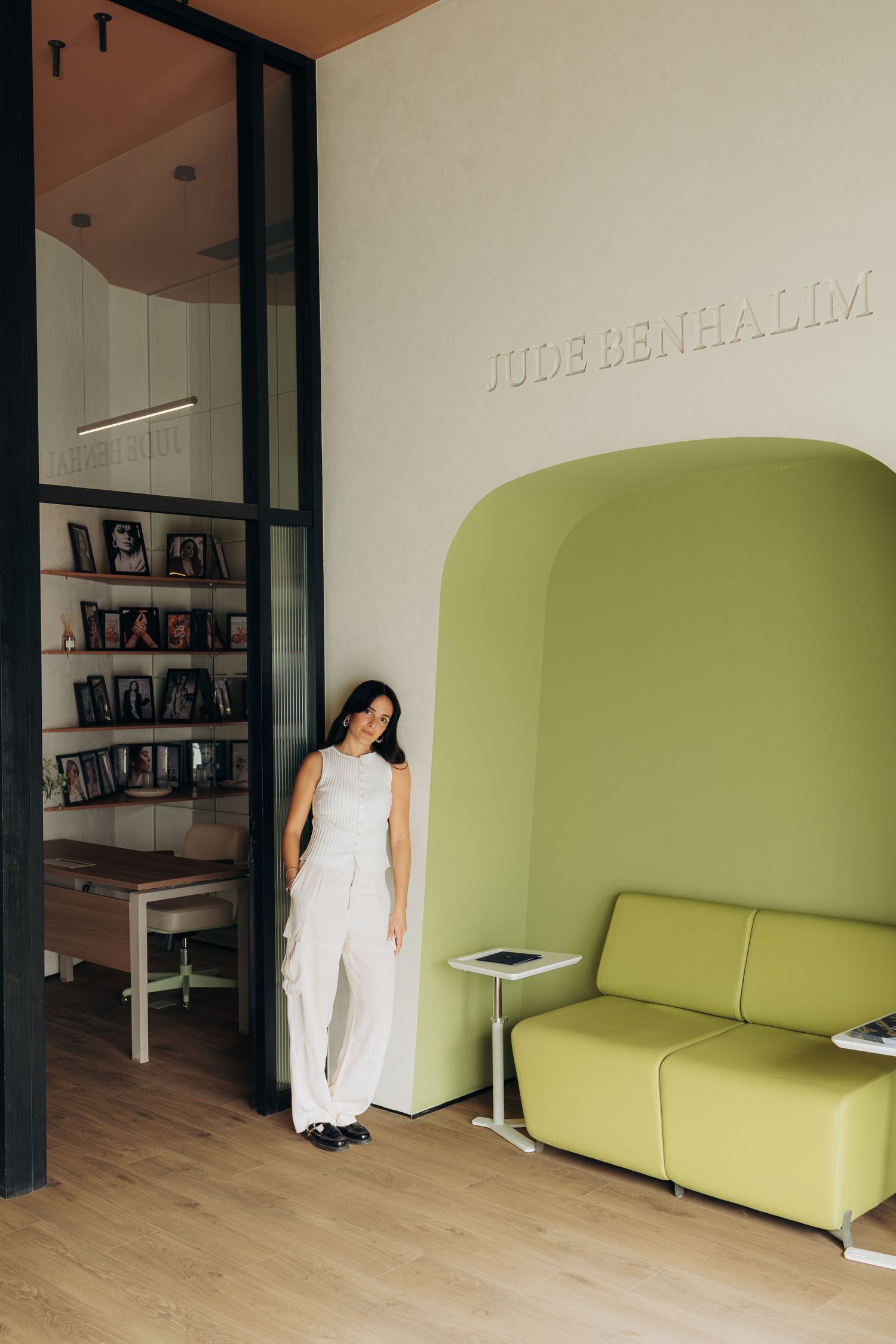 Woman in white outfit standing in modern office lobby with green arched sofa and Jude Benhalim branding on wall.