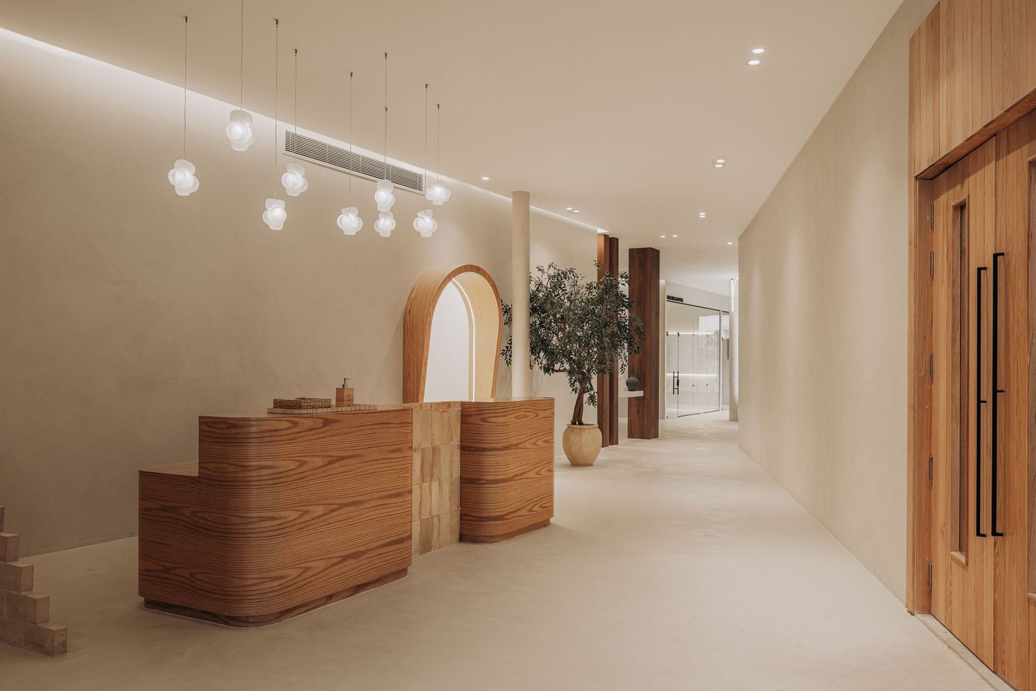 Elegant reception corridor with curved wood desk, globe pendant lights, arched mirror, and indoor olive tree.